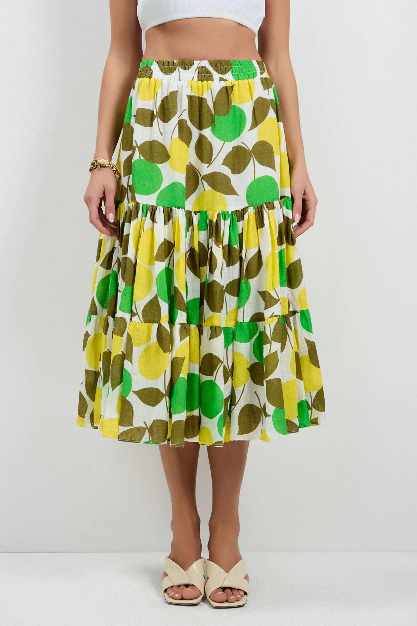 A woman wears the Marsay Drop Waist Midi Skirt, featuring a green, yellow, and white leaf print, an elastic waistband, and a flowy tiered midi design. She pairs it with beige sandals; only her lower body is shown.