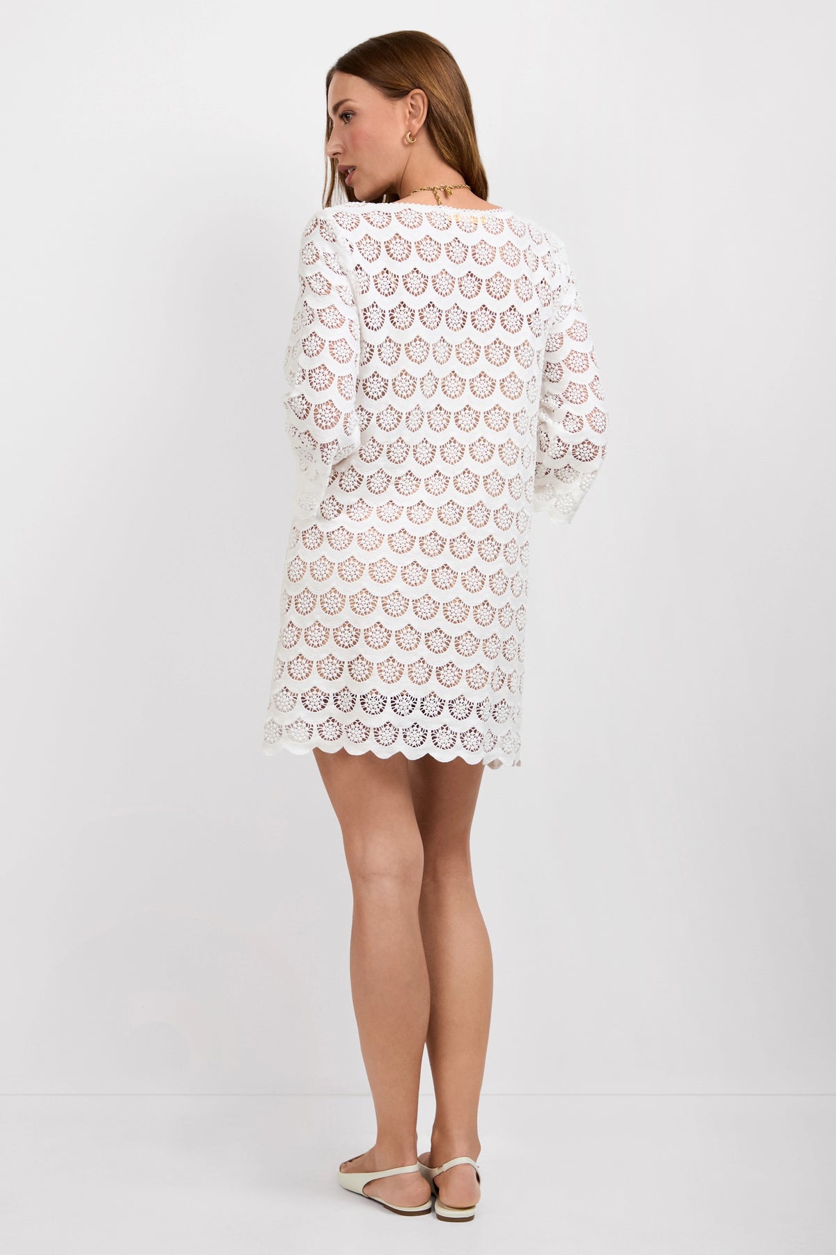 A woman with straight, light brown hair wears the Lita Crochet 3/4-Sleeve Dress in white, styled with flat white sandals. The knee-length dress features a scalloped crochet pattern and is shown against a plain white background.