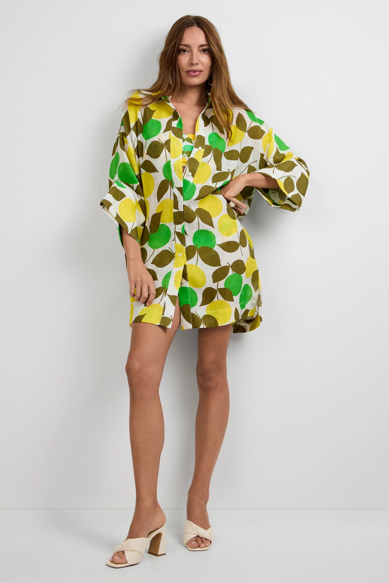 A woman with long brown hair stands against a white wall wearing the Opponent Shirt Dress featuring a bold green, yellow, and brown leaf print, paired with cream heeled sandals.