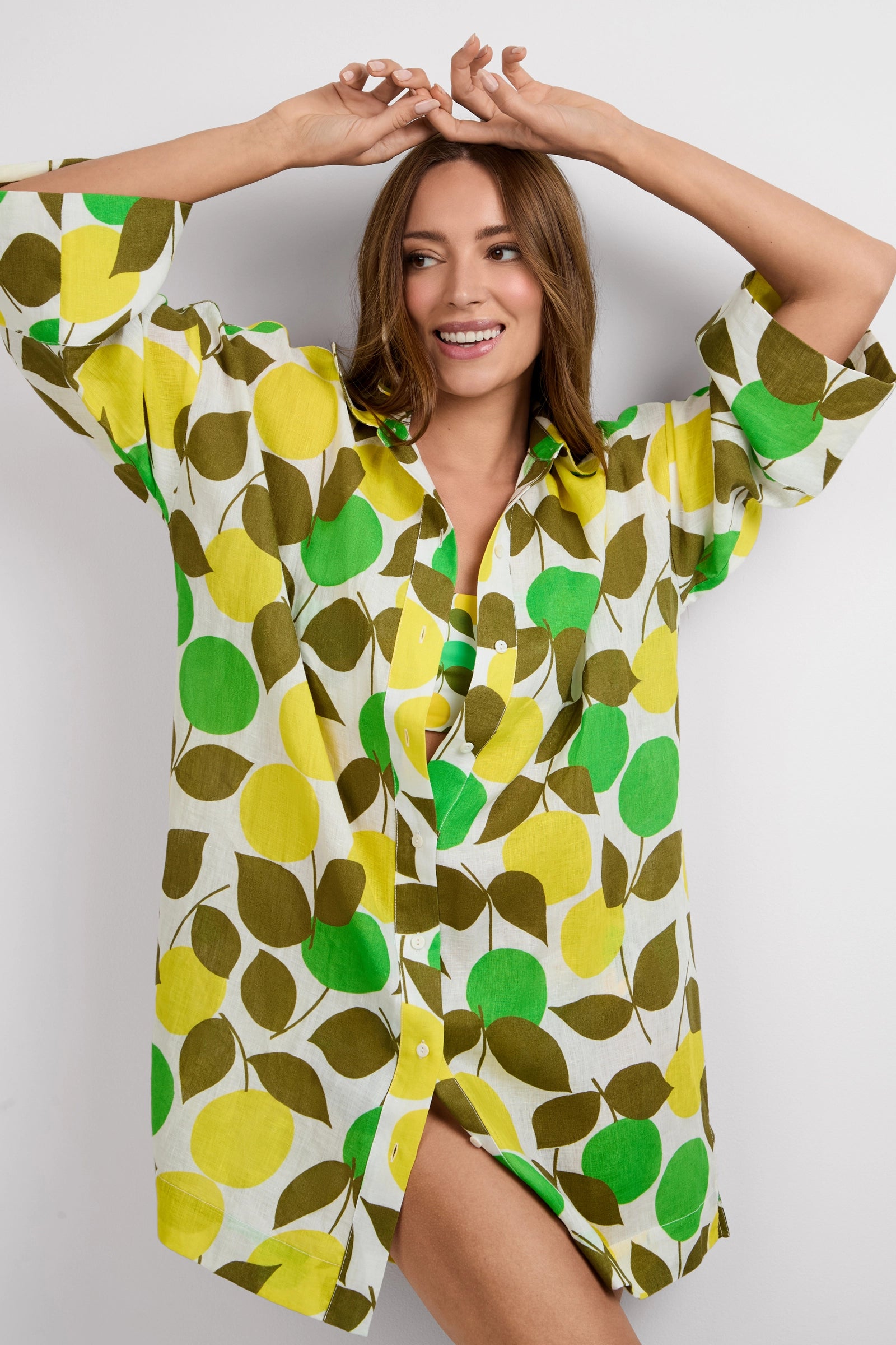 A woman smiles with her hands raised above her head, wearing the Opponent Shirt Dress, a loose button-up featuring a green, yellow, and brown apple and leaf pattern on a white background.