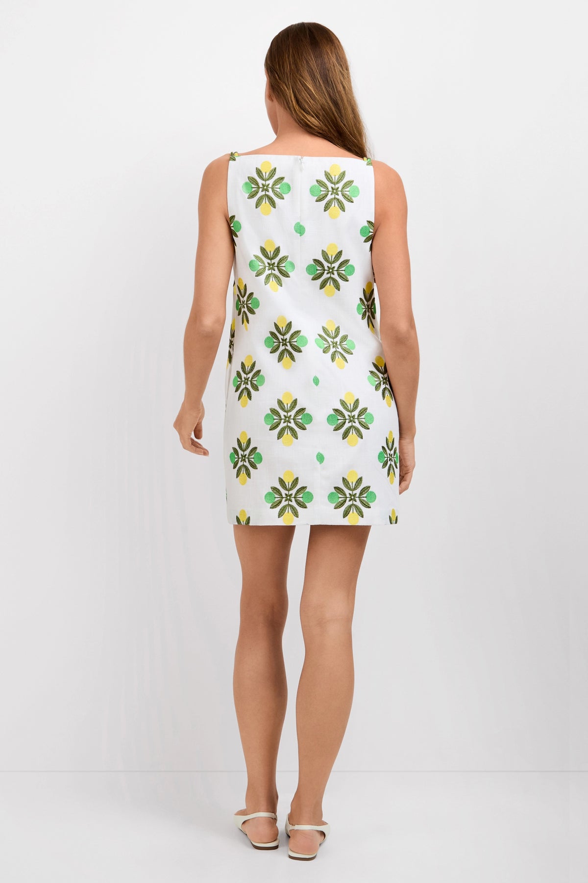 A woman with long, straight hair stands against a plain white background, facing away and wearing the Solay Mini Dress—a sleeveless white mini dress with green and yellow floral patterns—paired with white flat shoes.