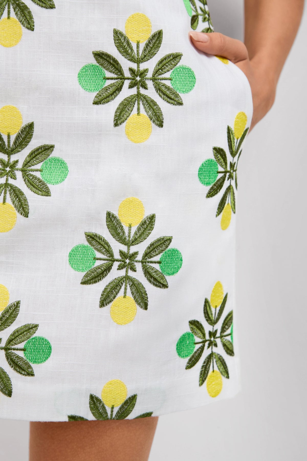 A close-up of the Solay Mini Dress, featuring white fabric with green and yellow lemon and leaf embroidery; a person’s hand is partially tucked into the side pocket.