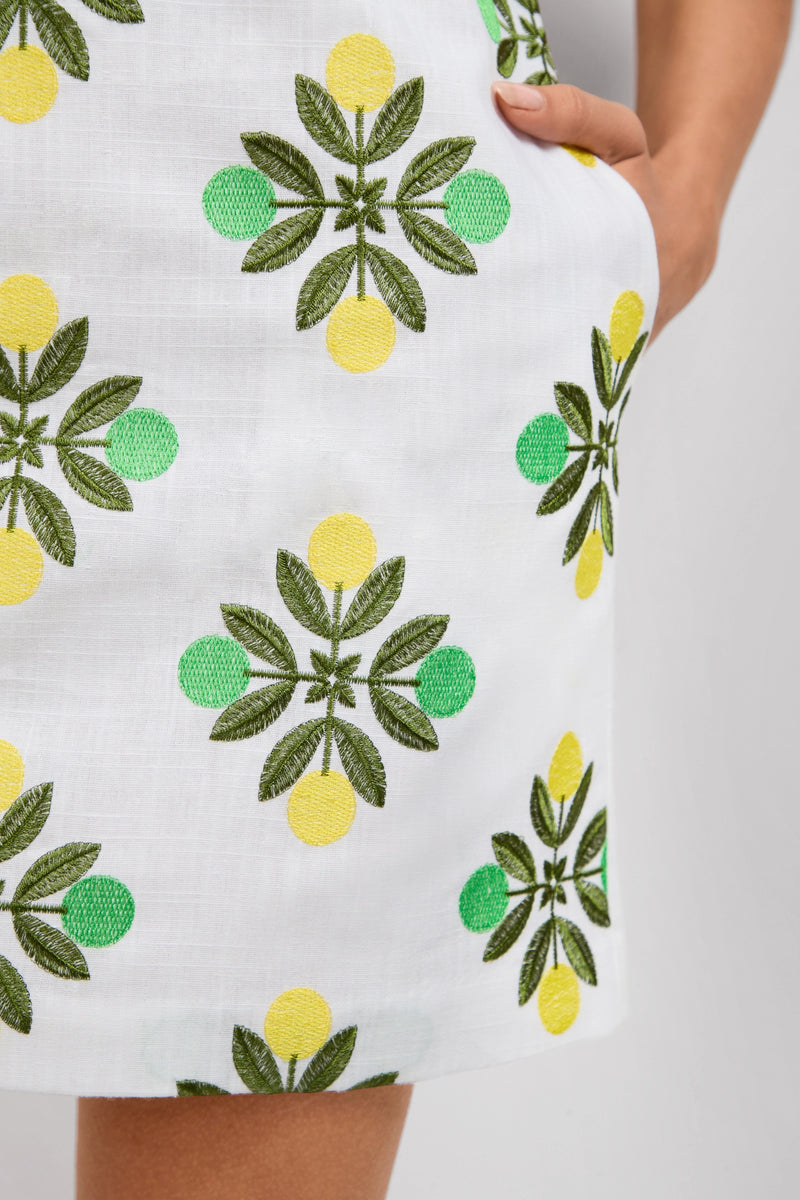 A close-up of the Solay Mini Dress, featuring white fabric with green and yellow lemon and leaf embroidery; a person’s hand is partially tucked into the side pocket.