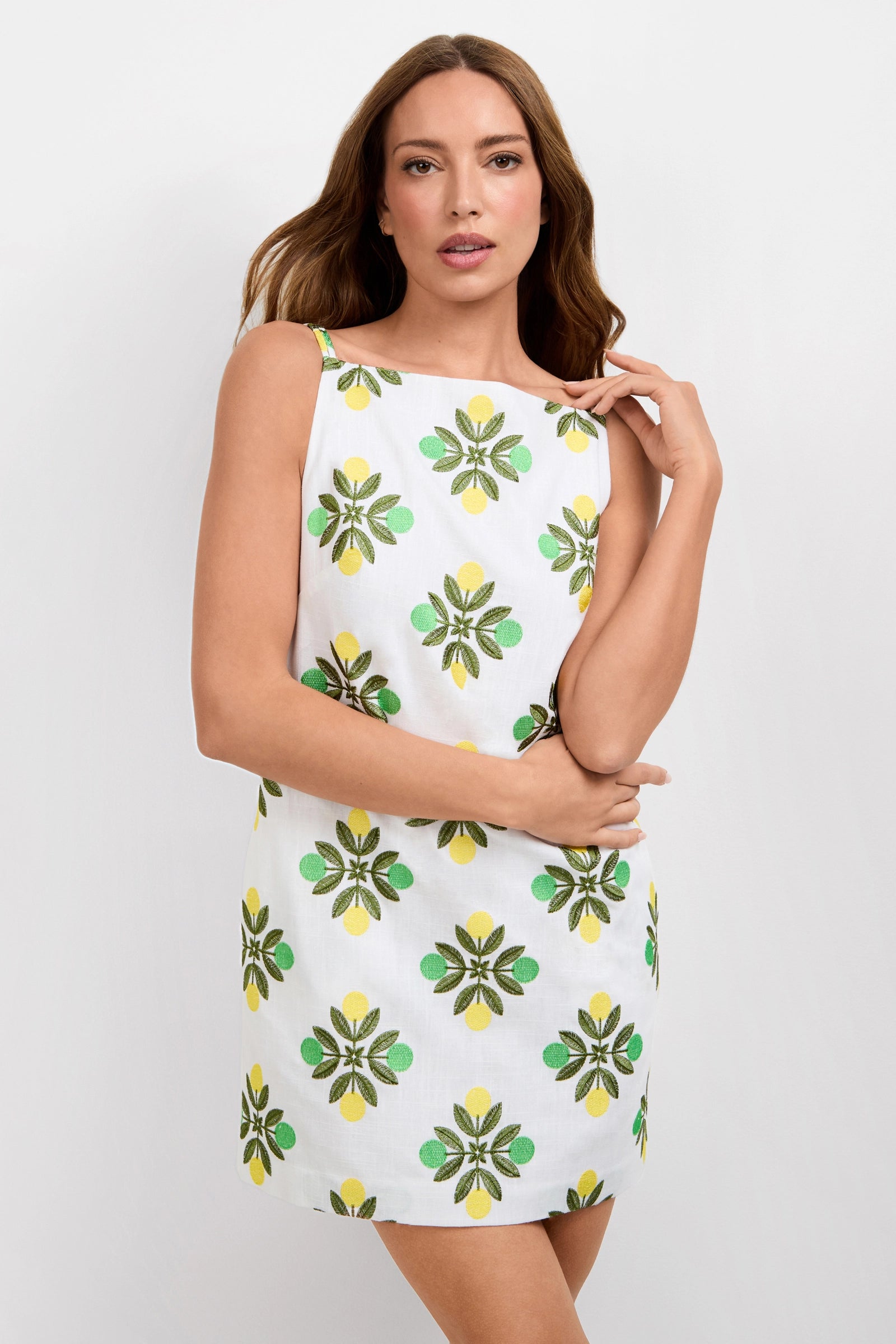 A woman models the Solay Mini Dress, a sleeveless white dress with green and yellow floral patterns, standing against a plain white background with one hand touching her hair.
