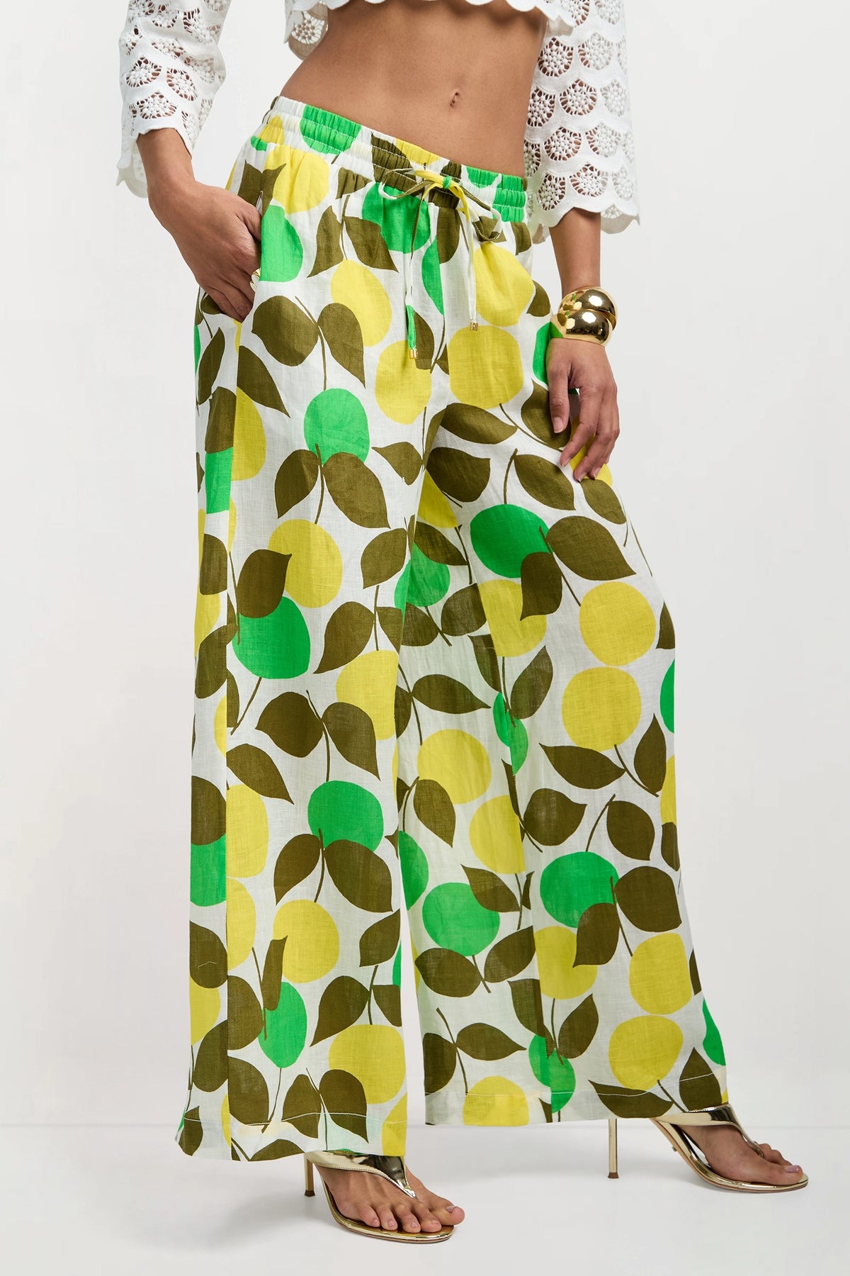 A woman models the Azule Wide-Leg Pants, featuring a green and yellow leaf print, styled with a white cropped eyelet top, gold heels, and chunky gold bracelets.