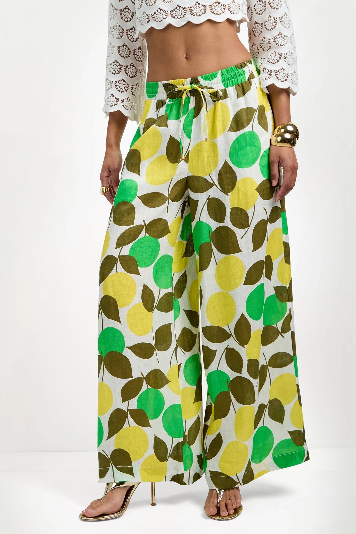 A woman wears Azule Wide-Leg Pants with a green and yellow leaf print, paired with a white crochet crop top, gold heeled sandals, and gold bangles.