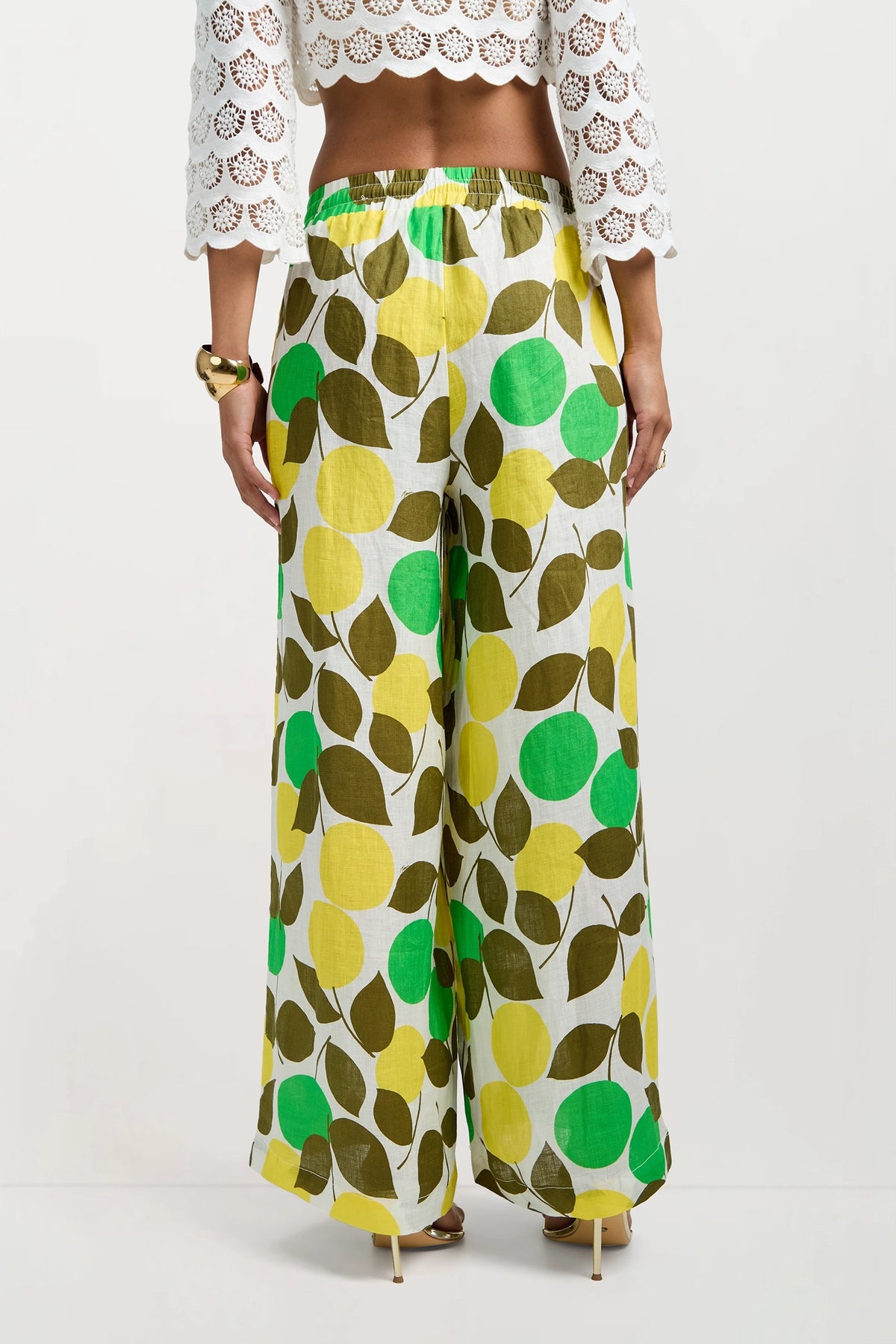 A woman wearing Azule Wide-Leg Pants with a green, yellow, and brown leaf and circle print, a white crochet cropped top with scalloped sleeves, high-heeled sandals, and a chunky bracelet, shown from the back.
