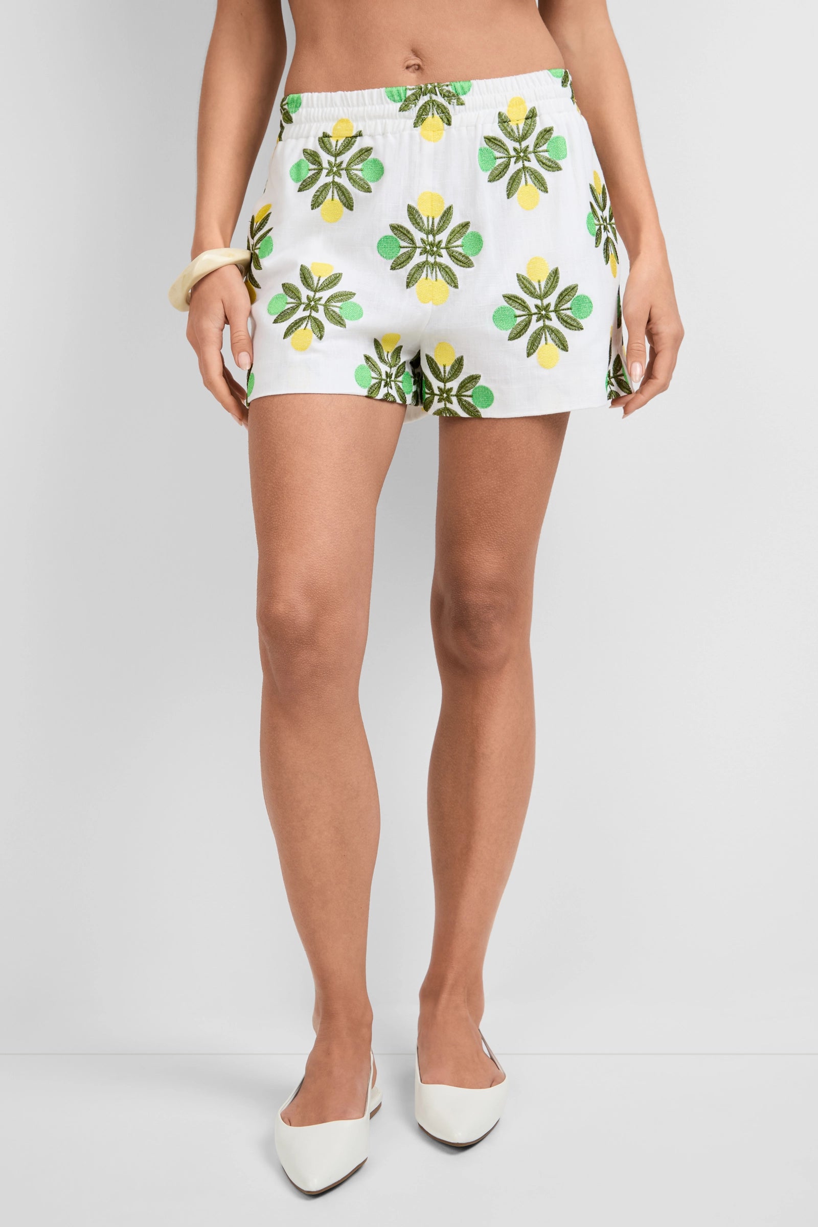 A person is wearing Azule Shorts with a green and yellow flower pattern, paired with a white bangle and white pointed-toe flats. The background is plain white, and only part of the person’s torso is visible.