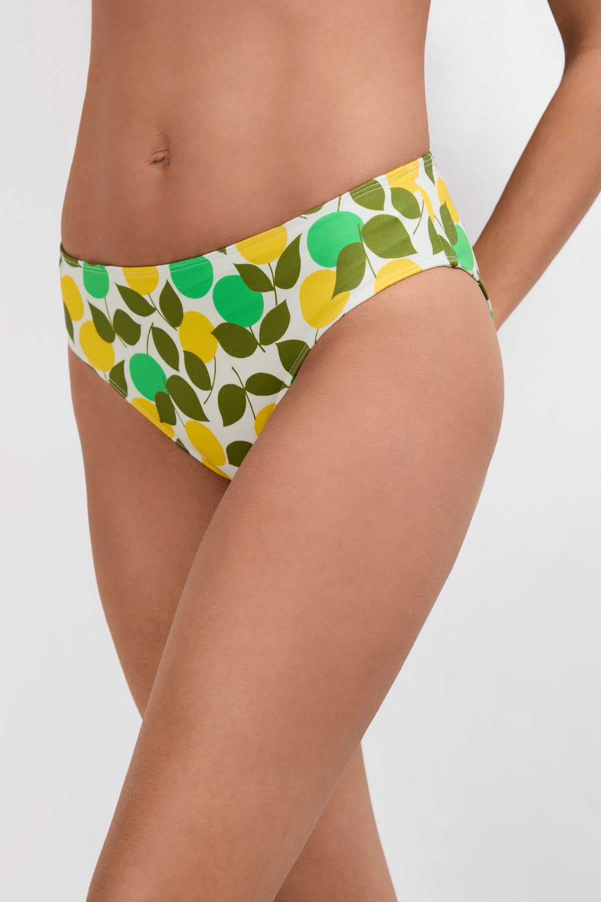 A person wears the Arissa Hipster Swim Bottom - Limone, featuring a green, yellow, and olive leaf pattern on white; only their torso and upper thighs are visible against a plain background.