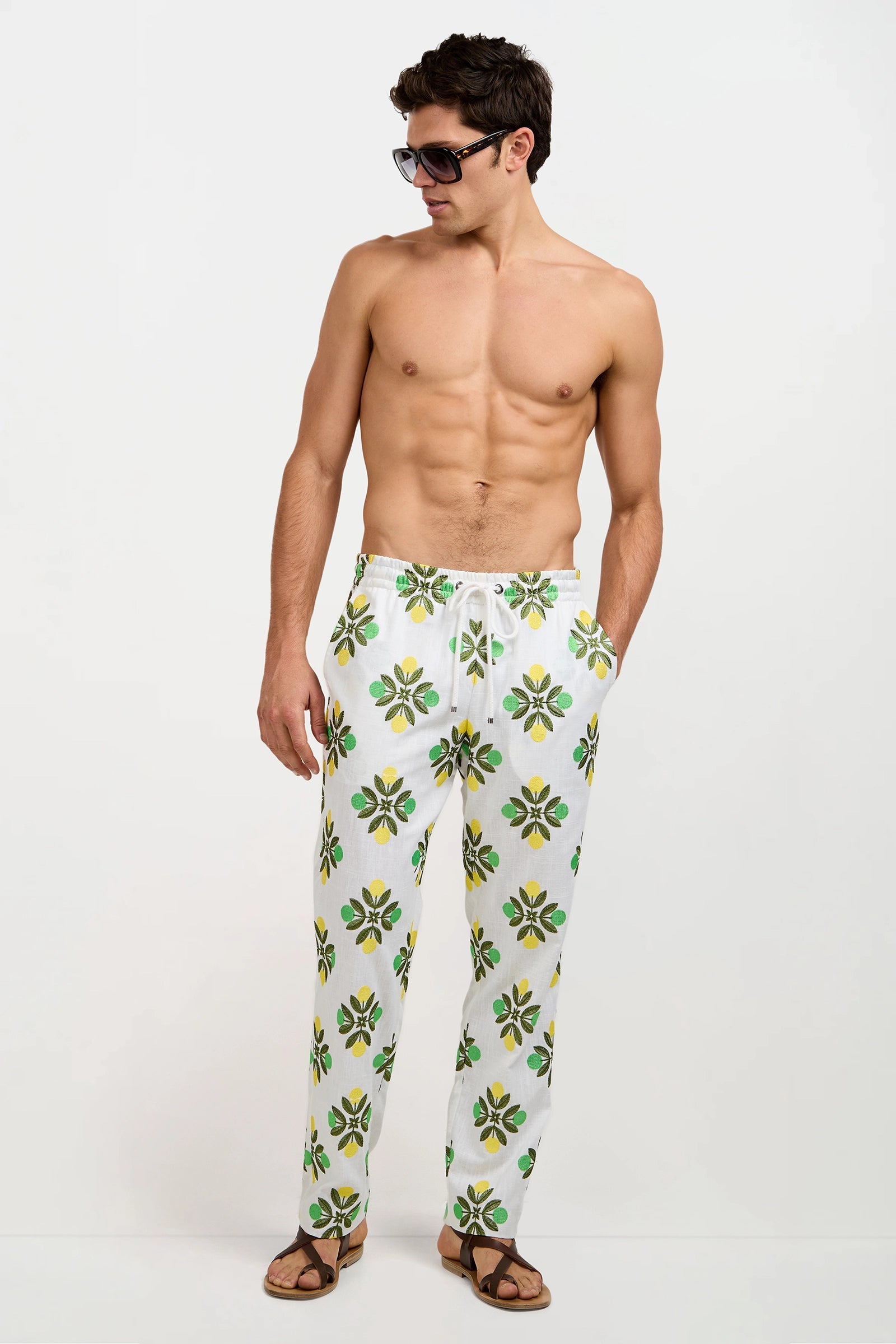 A shirtless man in sunglasses, brown sandals, and Felipe Drawstring Pant—white pants with a green and yellow floral print—stands against a plain white background, looking to his left.