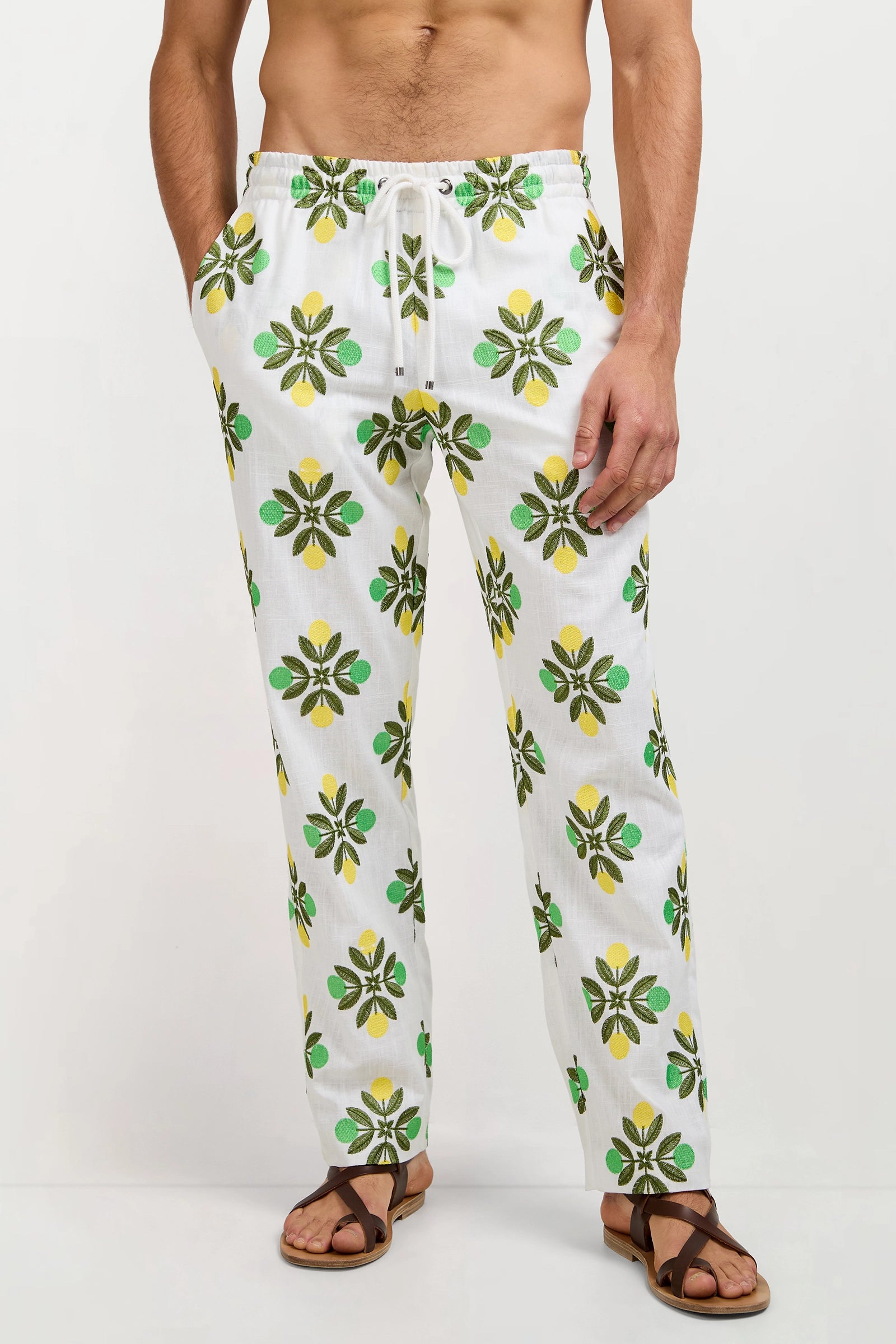 A man, hands in pockets, wears the Felipe Drawstring Pant featuring a yellow and green floral pattern, paired with brown sandals, standing against a plain background.