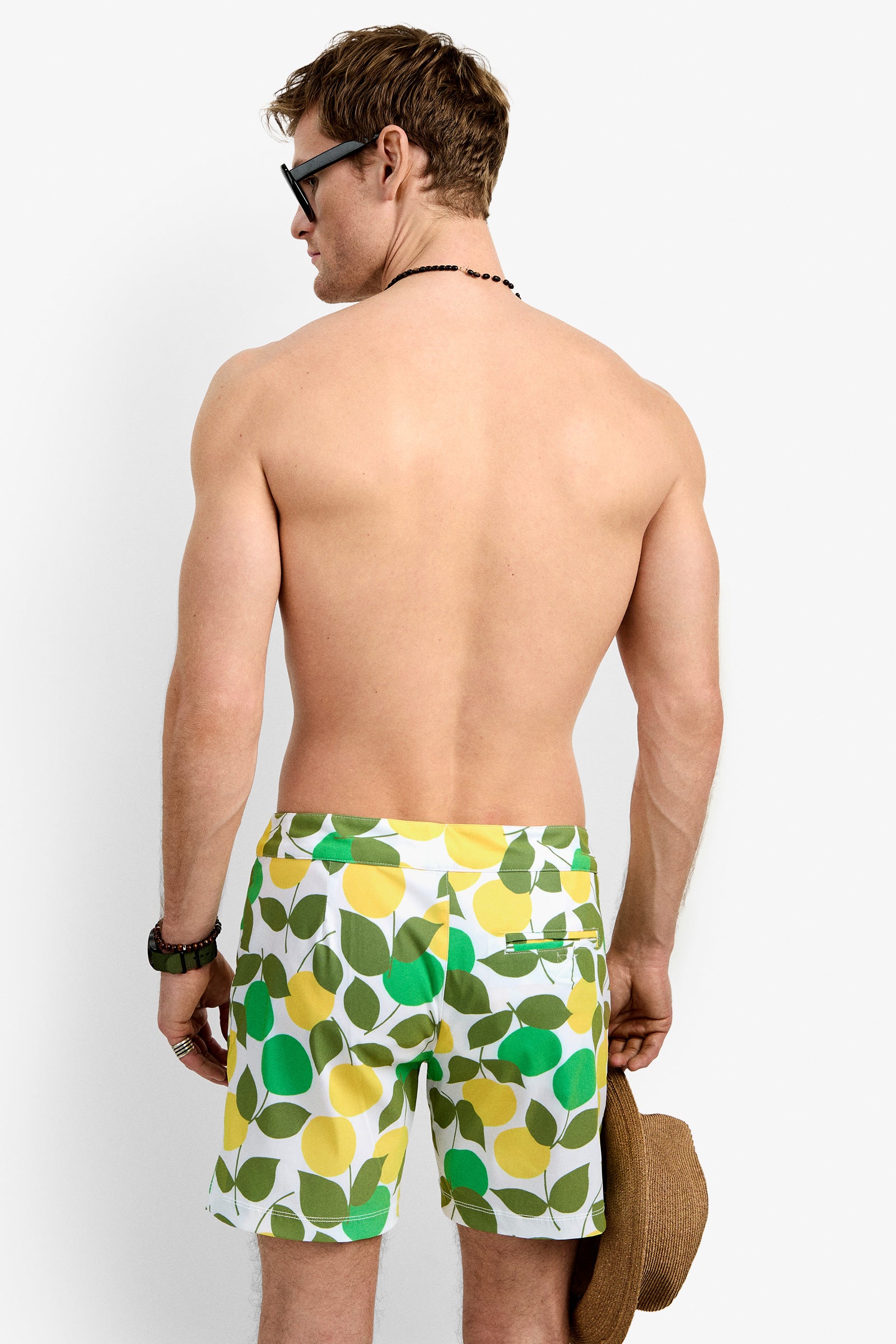 A man wearing Honolulu Board Short swim trunks with green, yellow, and white leaf patterns stands with his back to the camera, sunglasses on, holding a straw hat in his right hand.