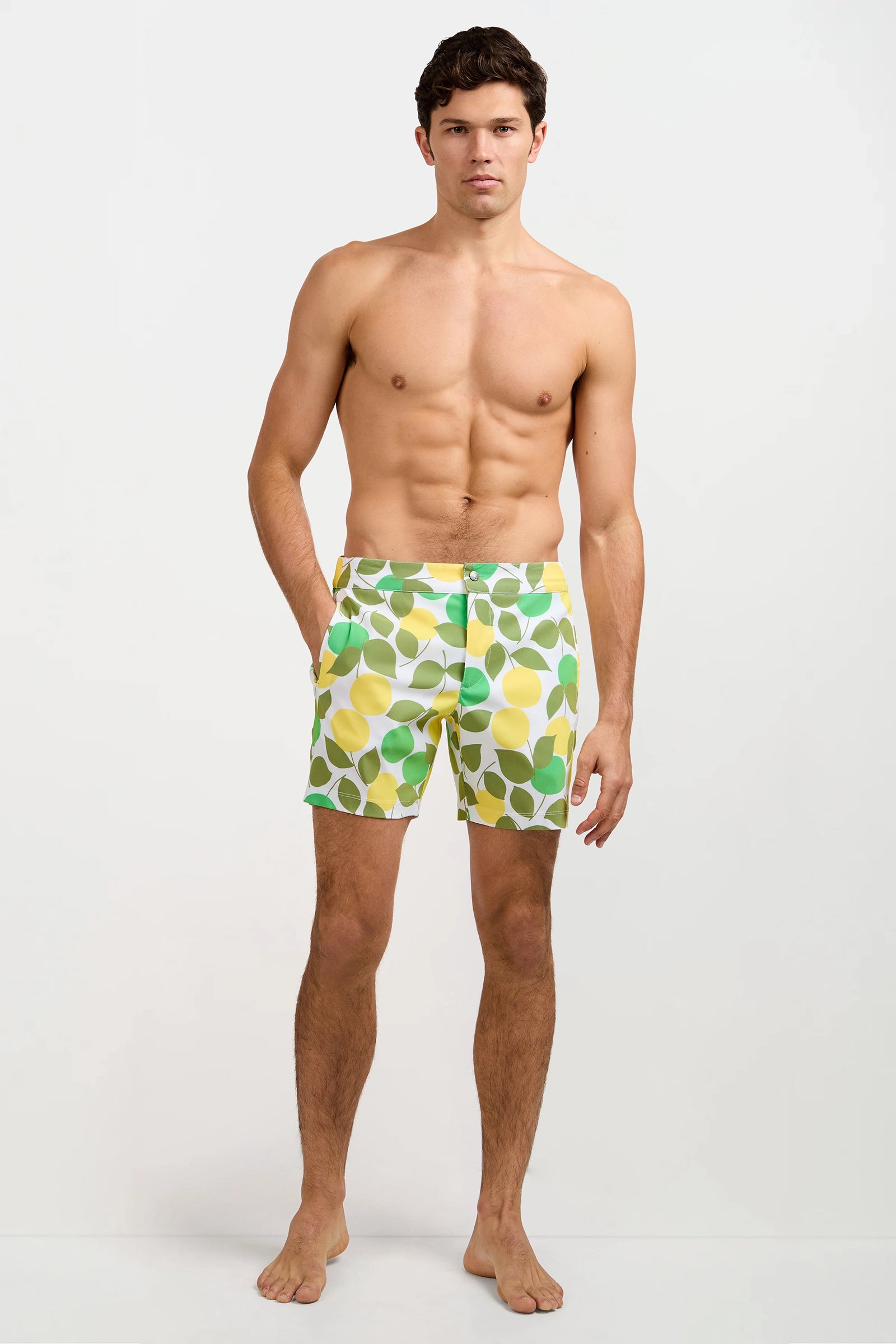 A man stands barefoot against a plain white background, wearing the Honolulu Board Short - Limone with a green and yellow lemon pattern. He is shirtless, facing forward with a neutral expression and his hands in his pockets.
