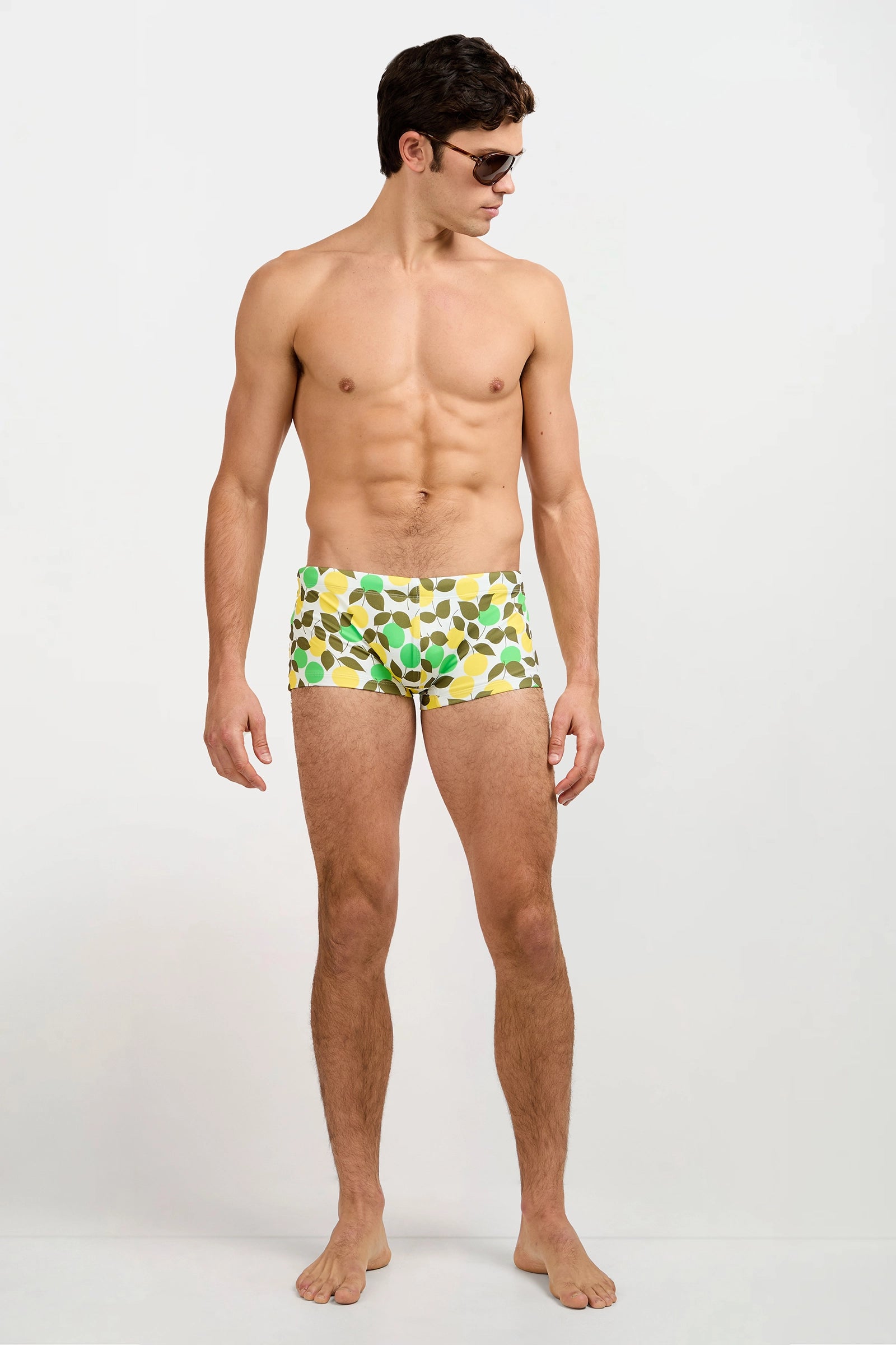 A man wearing the Sorrento Swim Trunk - Limone, featuring a green and white leaf pattern, stands barefoot against a plain white background, looking to his left while wearing sunglasses.