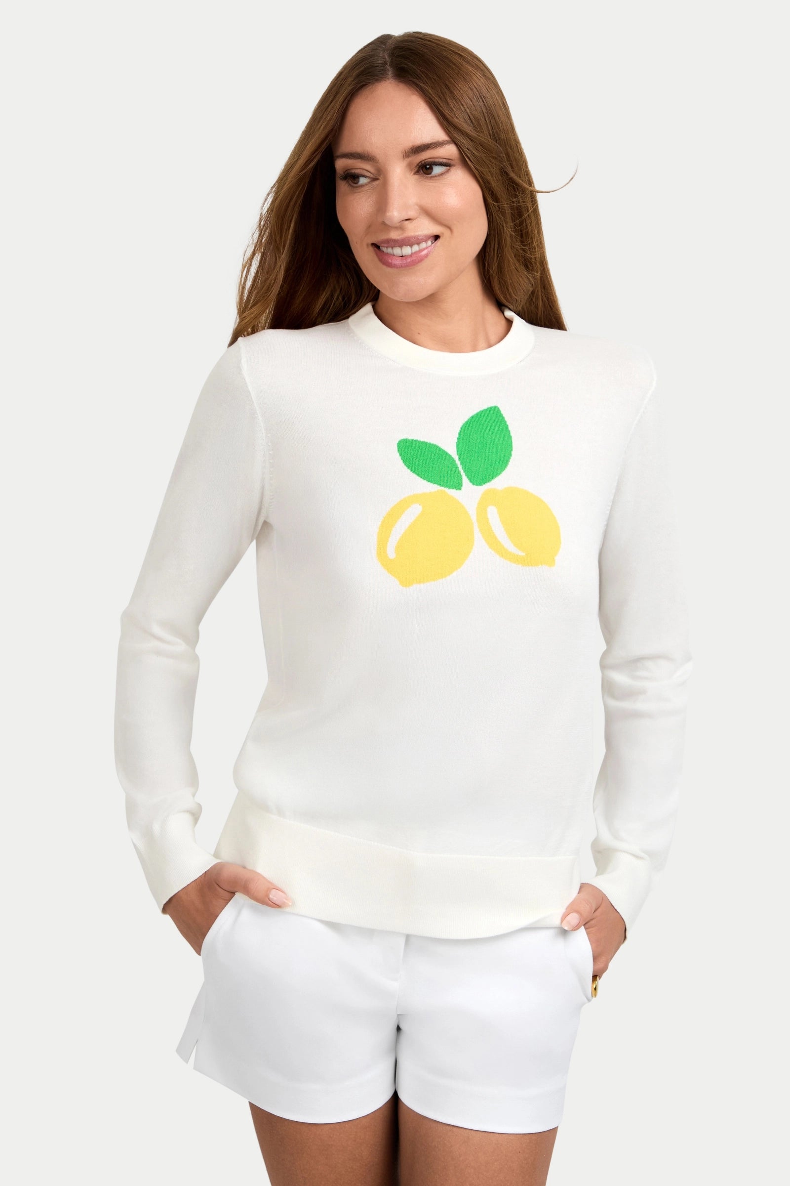 A woman smiles to the side with her hands in her pockets, wearing the Old Fashioned Sweater—a white long-sleeve top featuring two yellow lemons with green leaves on the front—paired with white shorts.
