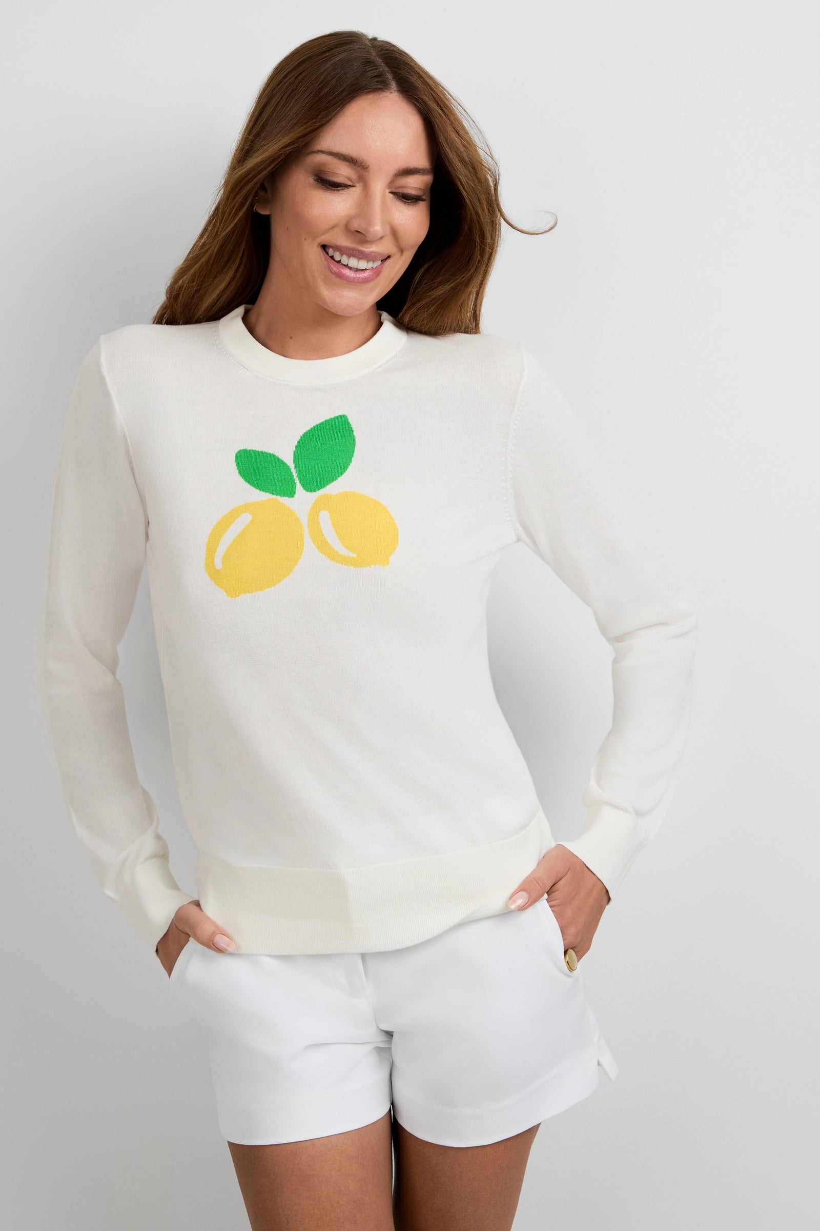 A woman with long brown hair smiles in the Old Fashioned Sweater, featuring two yellow lemons and green leaves on white, paired with white shorts. She poses against a plain white background.