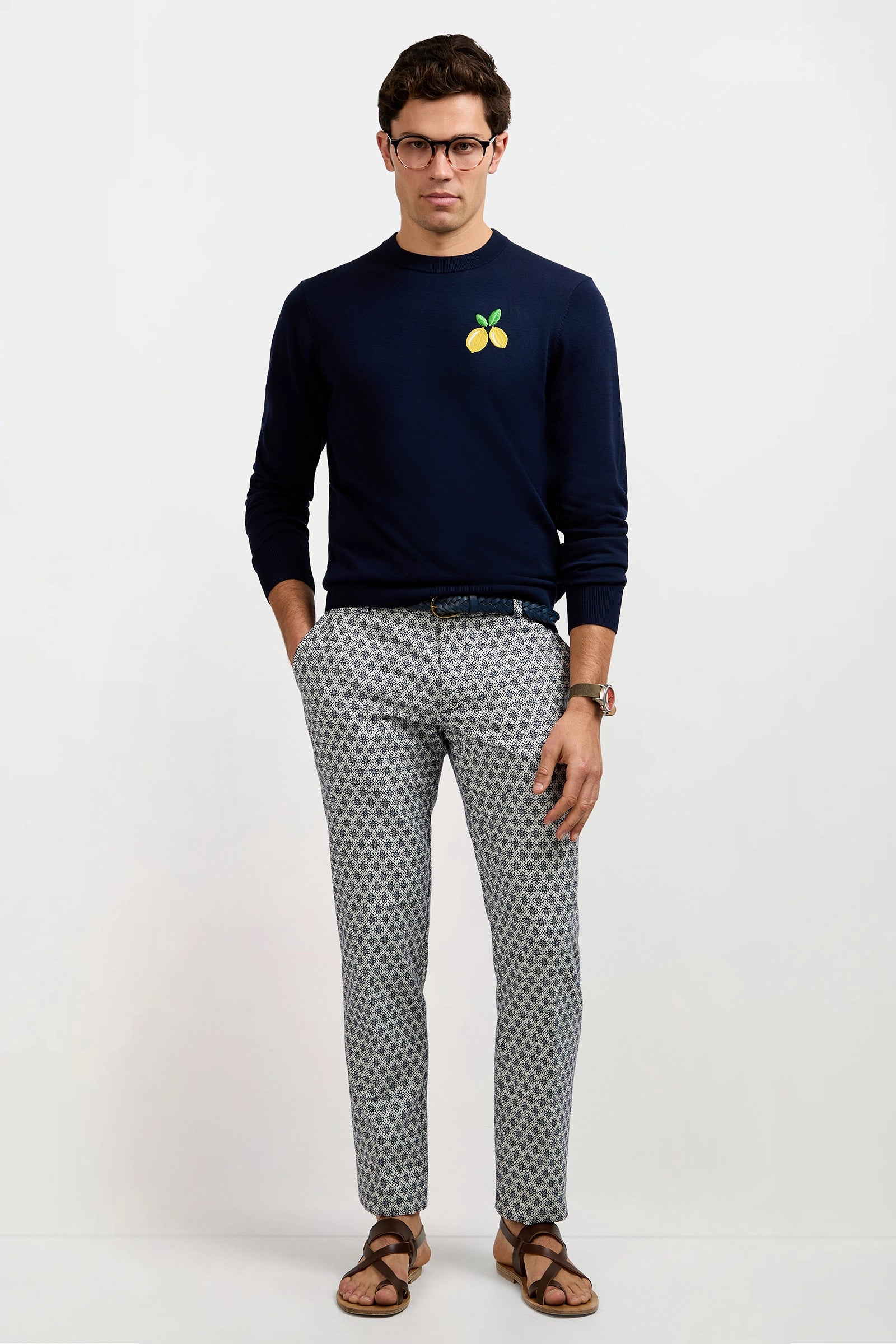 A man wears the Clyde Slim Trouser - Daisy Jacquard, paired with a navy lemon graphic sweater, glasses, a watch, and brown sandals, standing against a plain white background.