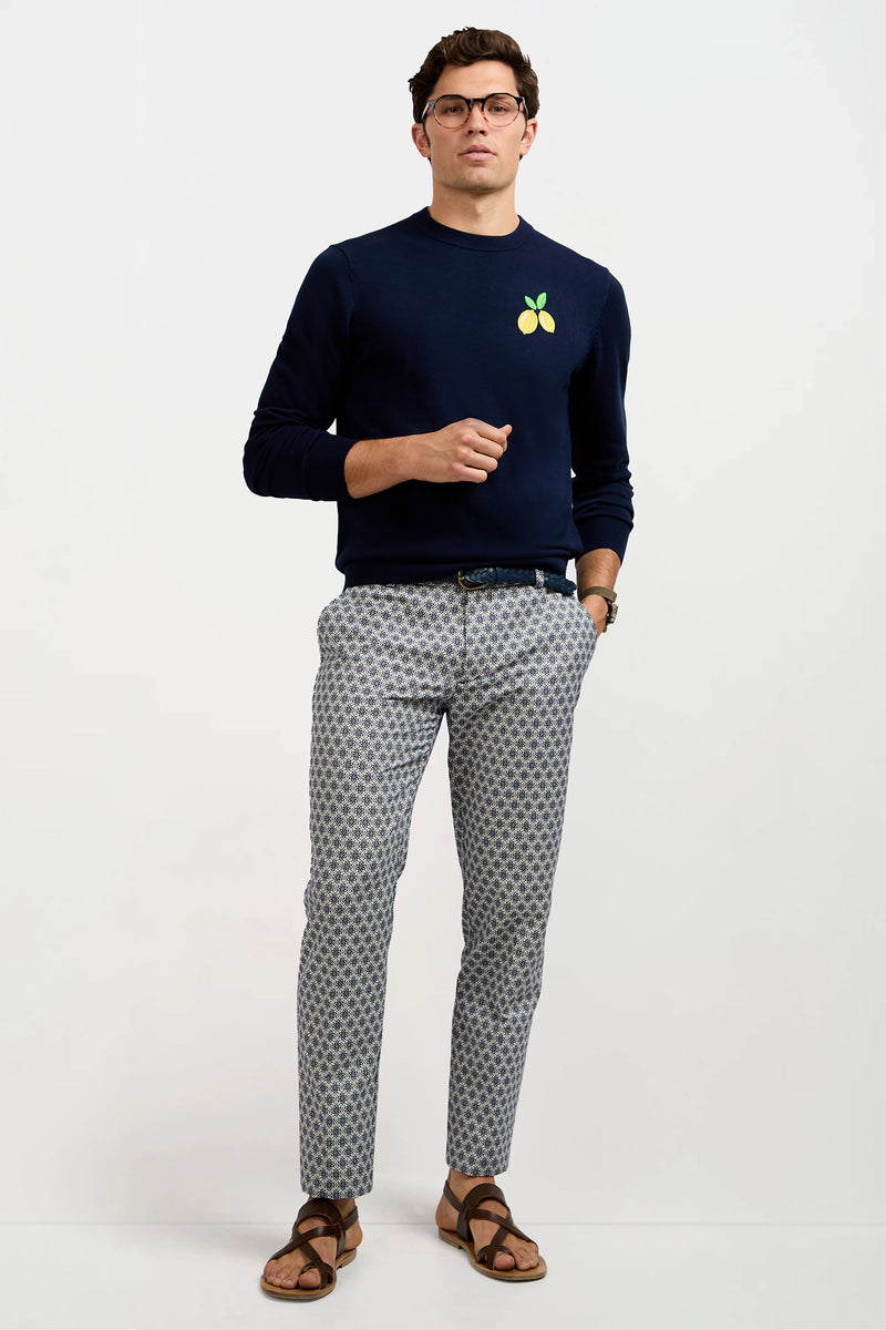 A man stands against a plain background wearing glasses, brown sandals, light grey patterned pants, and the Meyer Long-Sleeve Crewneck Sweater in navy with a small lemon graphic. One hand is in his pocket; the other holds his sweater.