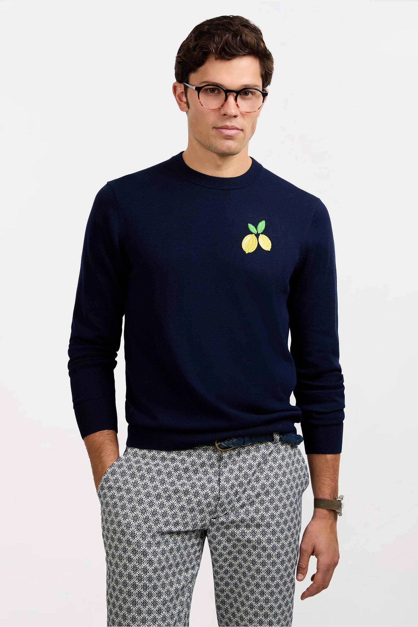 A man stands against a plain light background with his hands in his pockets, wearing glasses and the Meyer Long-Sleeve Crewneck Sweater in navy blue, which features a small lemon graphic on the left chest, paired with patterned pants.