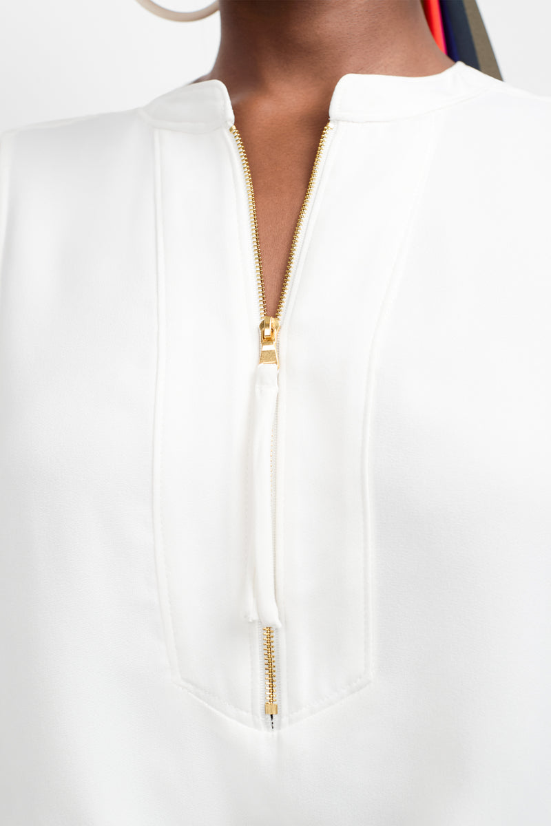 A close-up of a person wearing the Sleeveless Kaiko Top, featuring a white fabric, gold zipper partially unzipped at the neckline, small collar, and vertical seam details. The person's skin is visible at the neck.