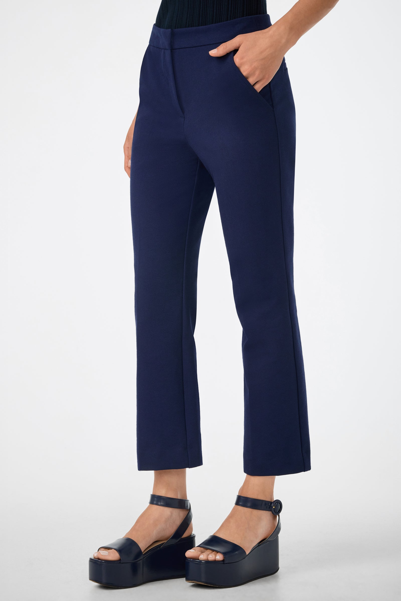 A person wearing the Banshee Pant—cropped navy blue dress pants—with hands in pockets and black platform sandals, standing against a plain white background.