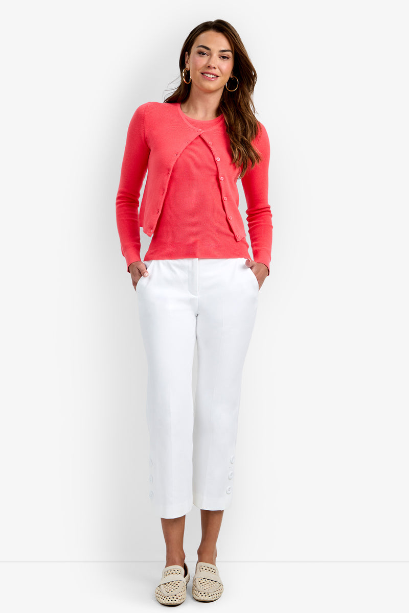 A woman stands with her hands in her pockets, wearing the Transcontinental Knit Cardigan in Corso Coral over a matching top, paired with white cropped pants featuring button details at the hem and light woven slip-on shoes.