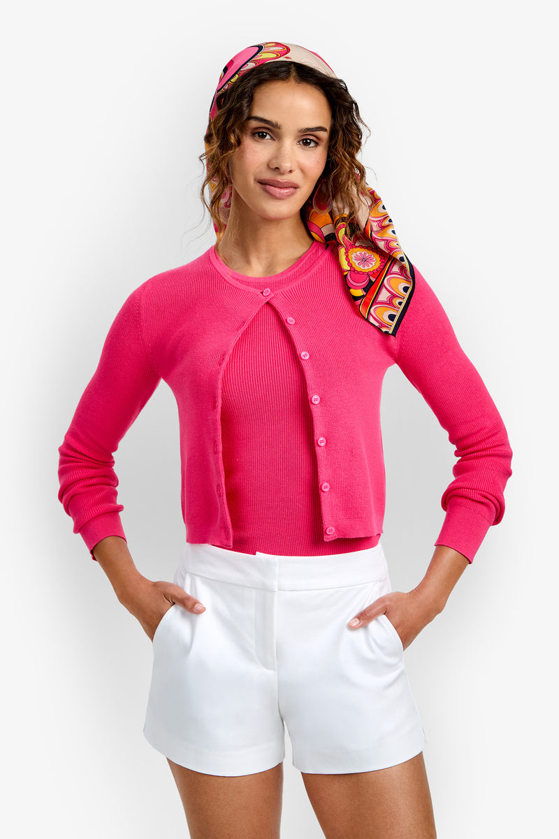 A woman wearing the Transcontinental Knit Cardigan in bright pink over a matching top, paired with white high-waisted shorts and a colorful patterned headscarf, stands with her hands on her hips against a plain white background.