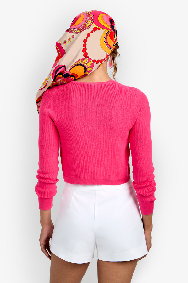 Woman seen from behind wearing the Transcontinental Knit Cardigan in pink, paired with white shorts and a colorful scarf featuring a vibrant circular pattern on her head, standing against a plain light background.