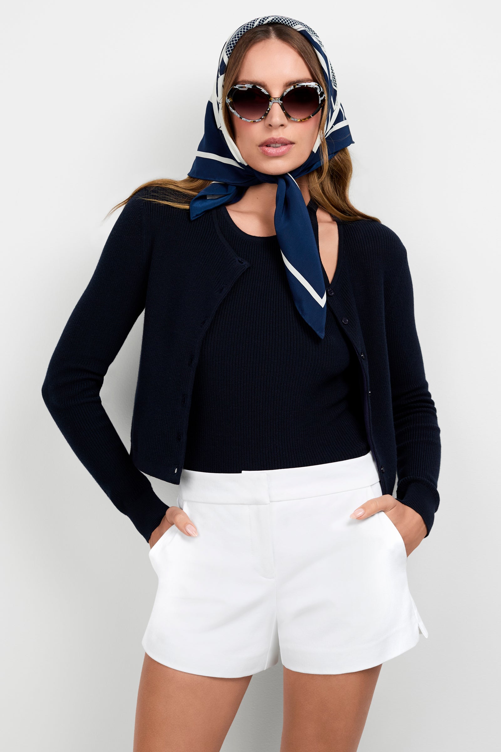 A woman models the Transcontinental Knit Cardigan in navy blue, paired with a matching top, white shorts, a navy and white headscarf, and round sunglasses, posing with hands in pockets against a plain white background.