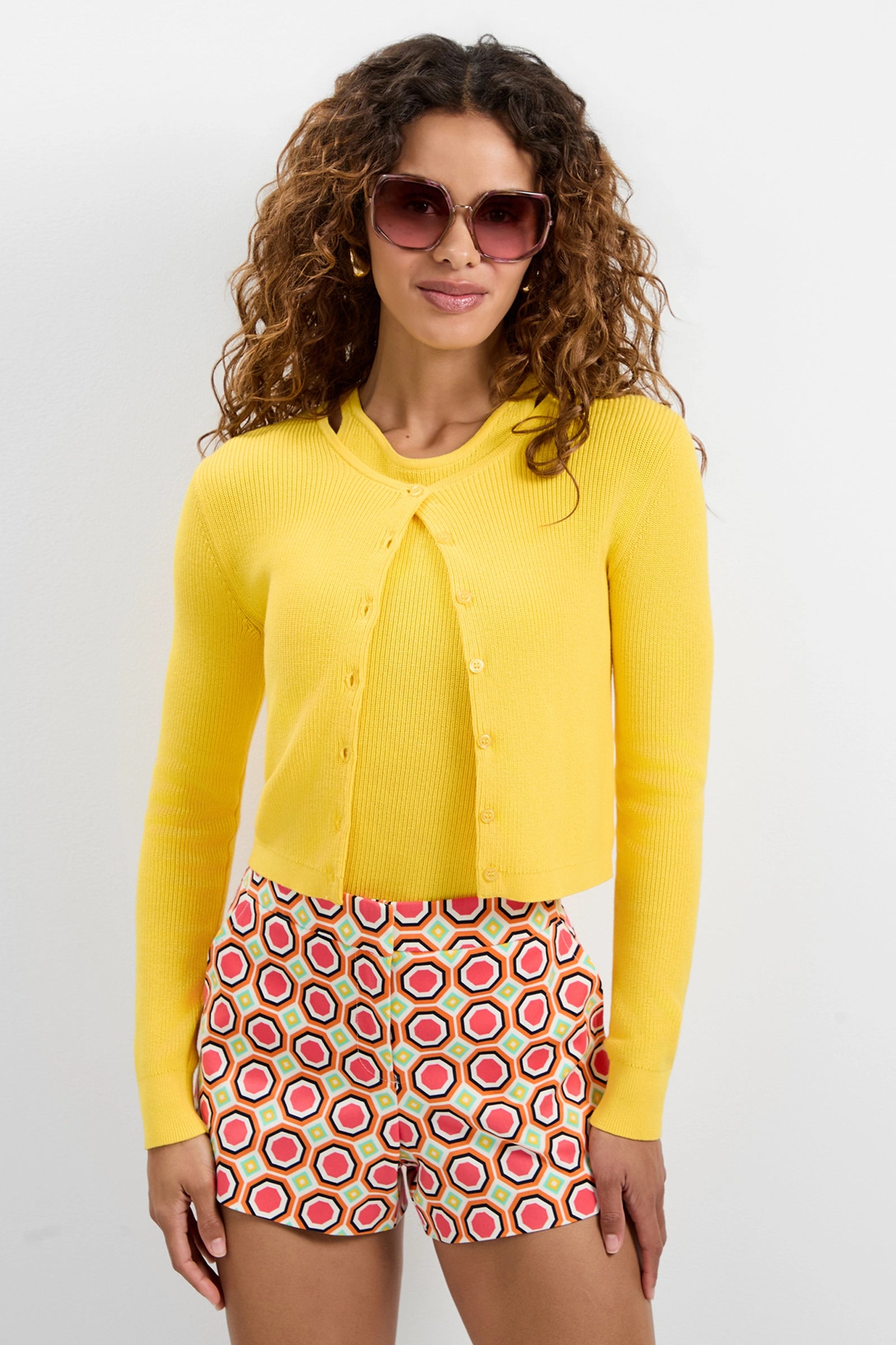 A woman with curly hair wears large sunglasses and the Transcontinental Knit Cardigan in yellow over a matching top, paired with colorful geometric shorts, standing against a plain white background and smiling slightly.