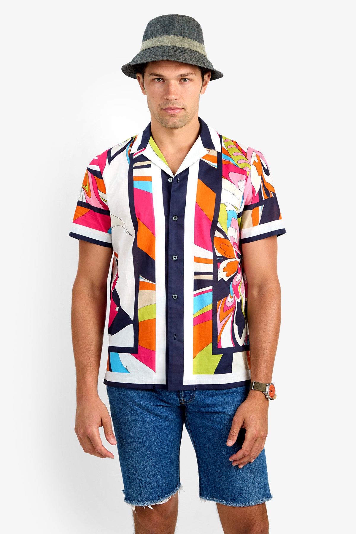 A man in a gray hat, the JOSUE CAMP SHIRT (100% cotton, colorful patterned short sleeves), blue denim shorts, and a watch stands facing the camera against a plain white background.