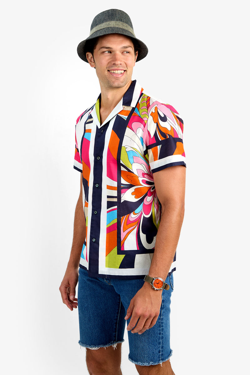 A man in a gray hat, blue denim shorts, an orange watch, and the JOSUE CAMP SHIRT—a colorful short-sleeve 100% cotton button-up with bold abstract patterns—smiles while looking left against a white background.