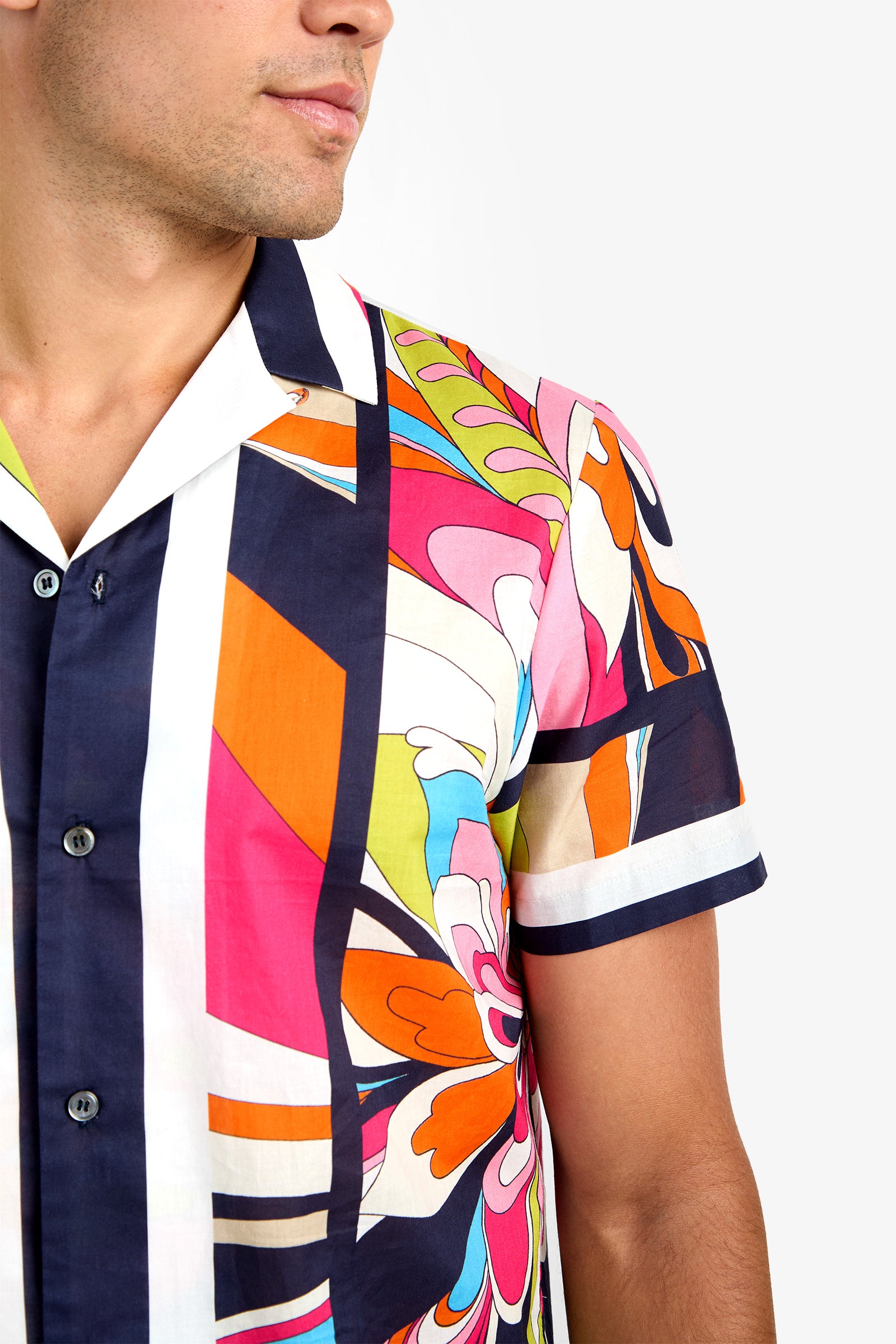 A man models the JOSUE CAMP SHIRT, a pure cotton, short-sleeve button-up with a bold abstract floral and geometric print in orange, pink, blue, green, and white. His upper body is pictured against a plain background.