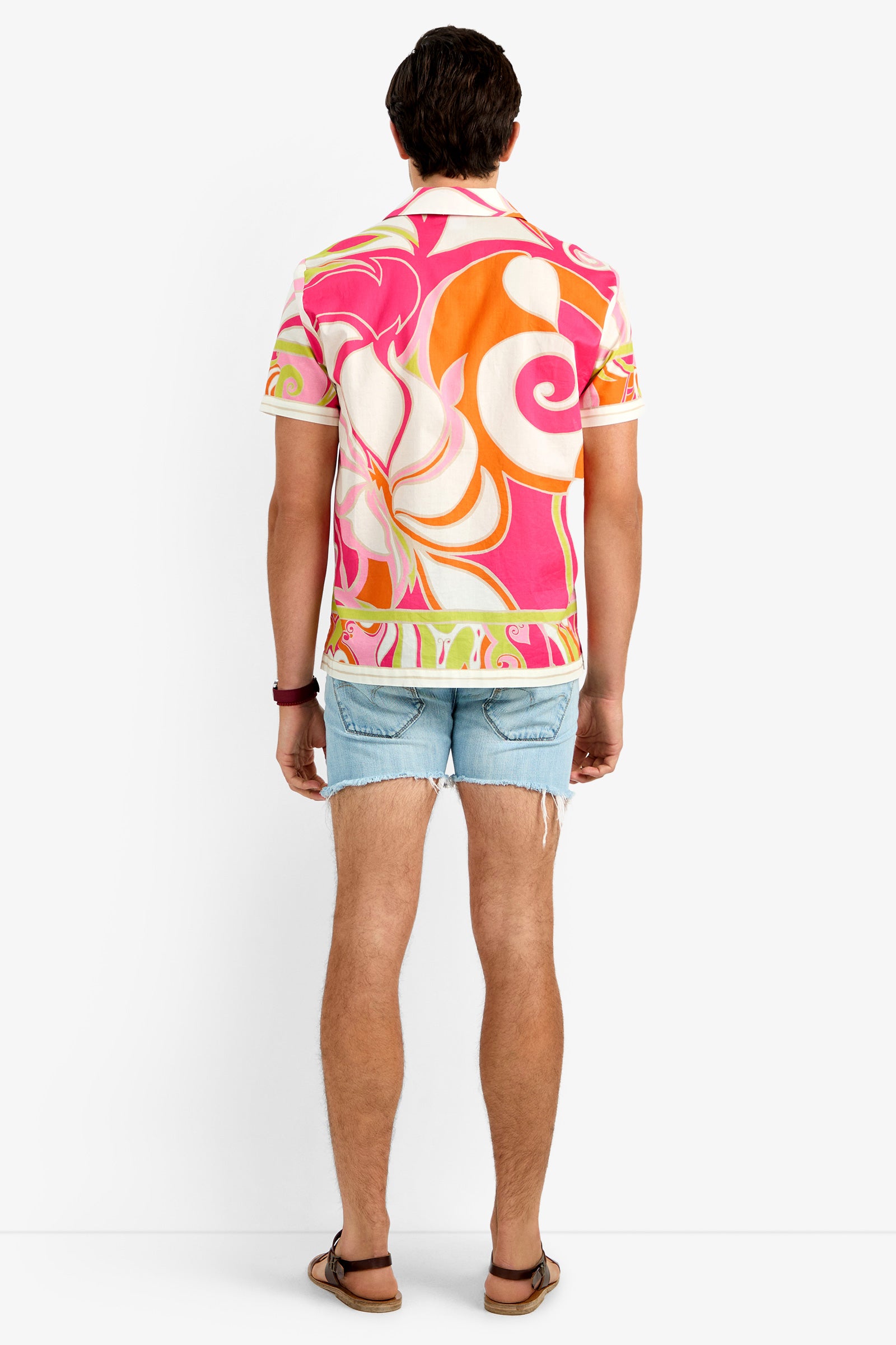 A person stands facing away in a JOSUE CAMP SHIRT with a pink, orange, and white pattern, light blue ripped denim shorts, brown sandals, and a red wristband against a plain white background.
