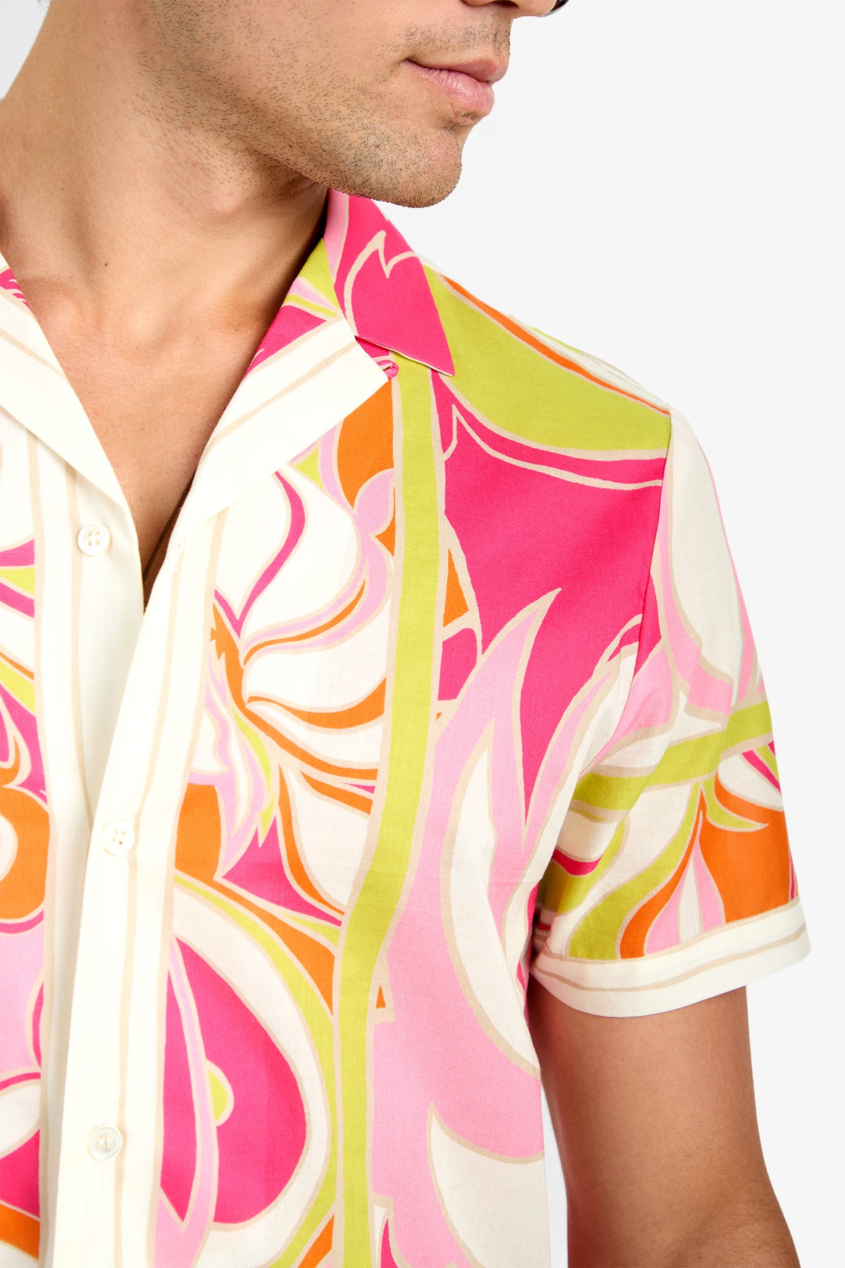 A person stands against a plain background wearing the JOSUE CAMP SHIRT, a short-sleeve cotton button-up featuring a bold and colorful pattern in pink, orange, lime green, and white. The person's face is partially visible.