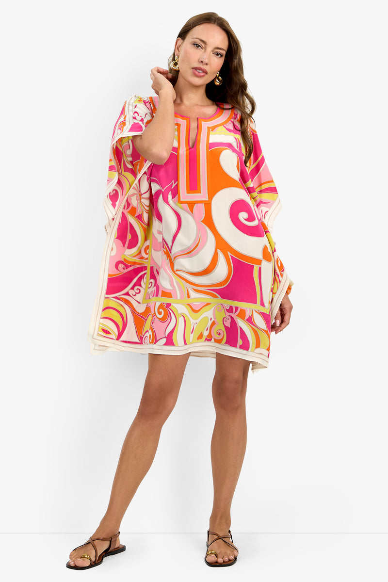A woman models the Theodora Caftan Dress, featuring a vibrant pattern in pink, orange, and white. She stands against a white background, posing with one hand in her hair and the other at her side, complemented by sandals and statement earrings.