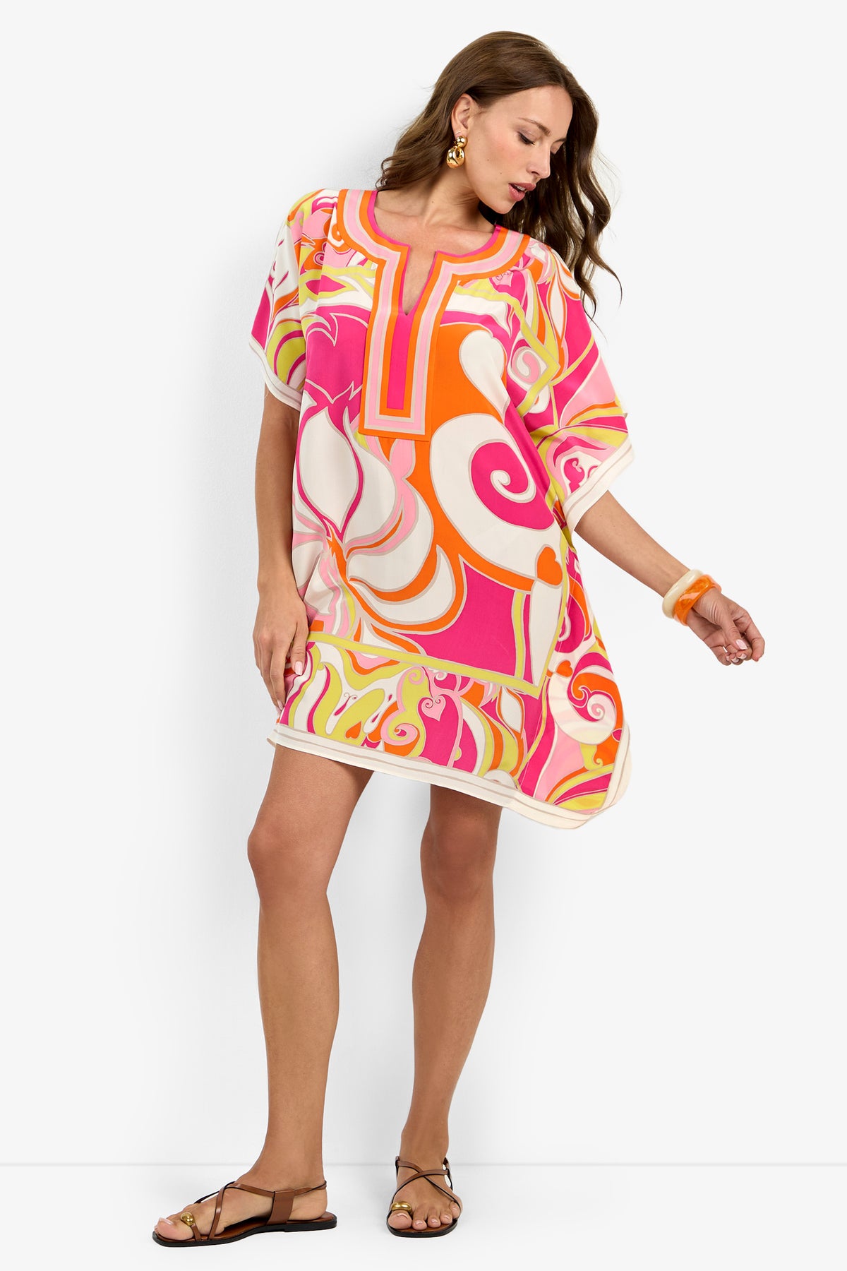 Model wearing the Theodora Caftan Dress in MULTI, featuring vibrant swirling patterns, paired with tan sandals, an orange bangle, and gold earrings against a white background.