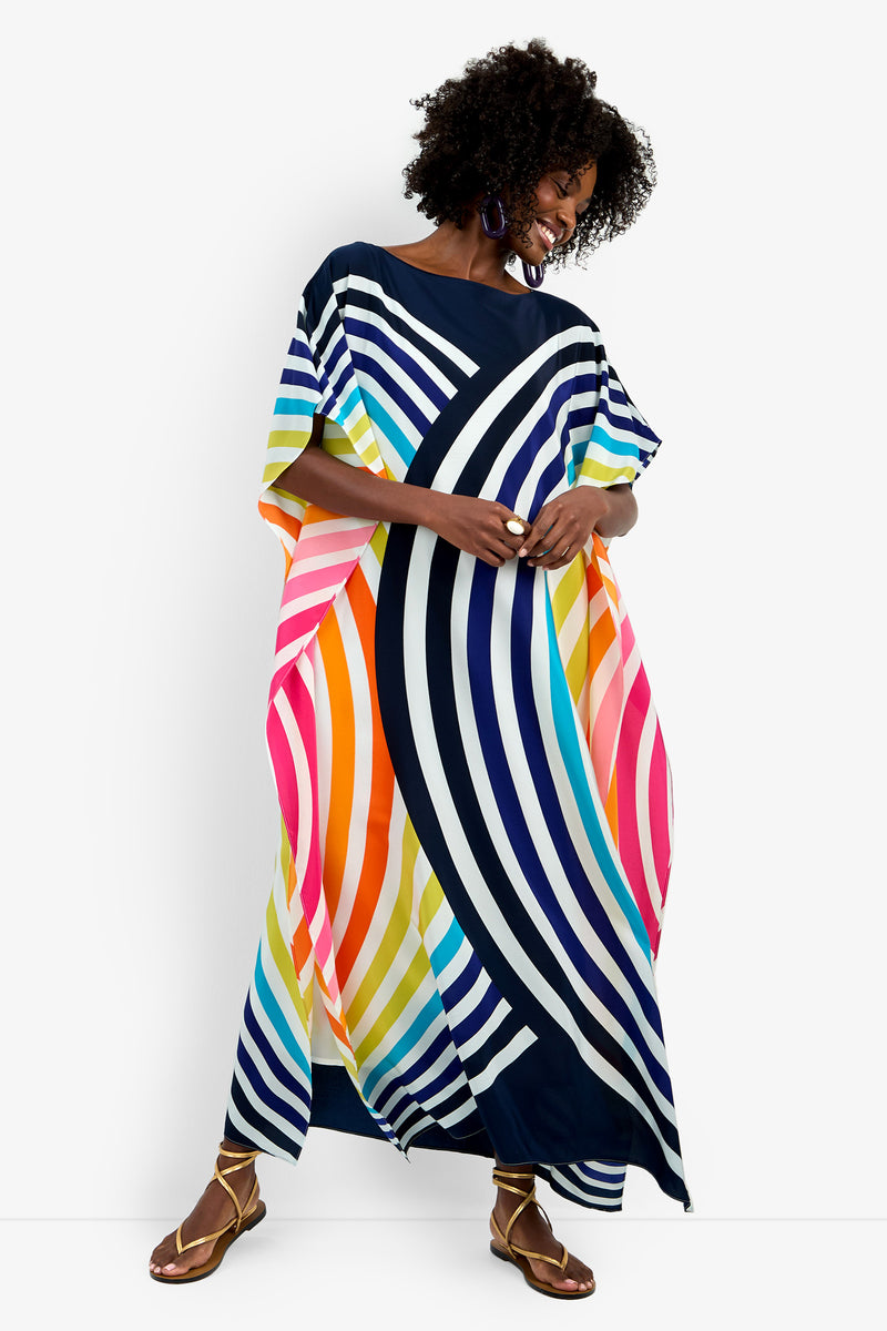 A woman smiles with her head tilted and hands clasped, wearing Theodora Caftan Maxi Dress—a vibrant, loose-fitting pure silk maxi with bold blue, pink, yellow, orange, and white diagonal and vertical stripes.