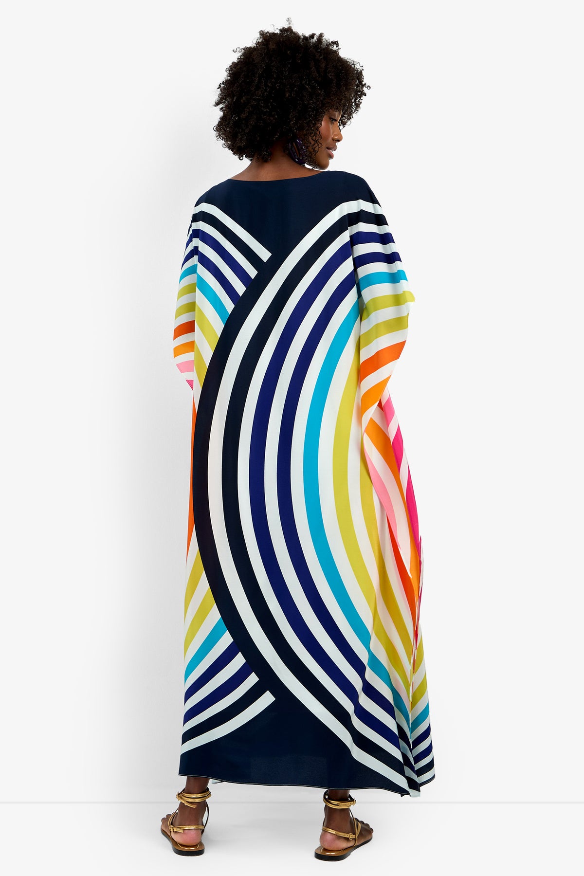 A woman with curly hair faces away, revealing the back of the Theodora Caftan Maxi Dress— a long, flowing pure silk dress with bold curved stripes in blue, yellow, orange, pink, and white—paired with sandals.