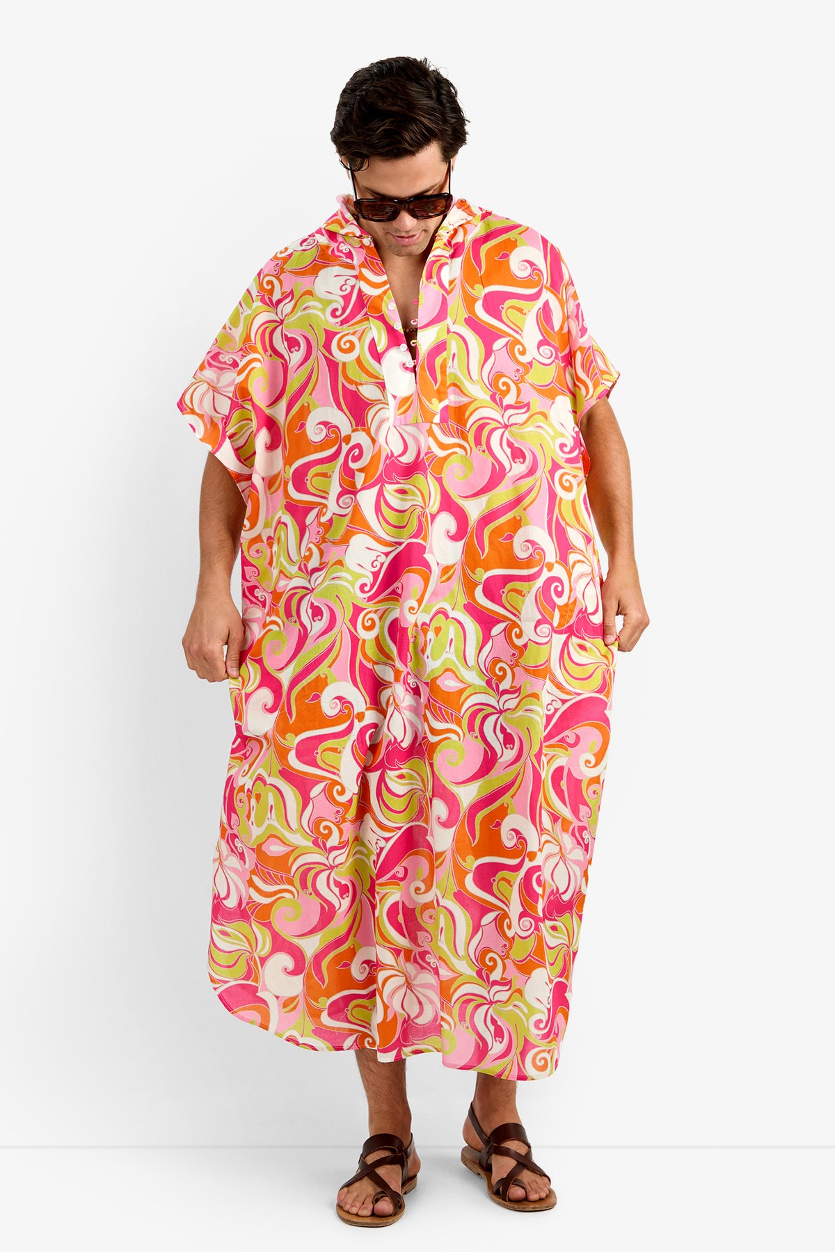 Wearing sunglasses and brown sandals, a person stands in the HURLEY CAFTAN—a 100% cotton, loose-fit dress with bold pink, orange, red, and white swirls. They look down while holding the sides of this vibrant garment.