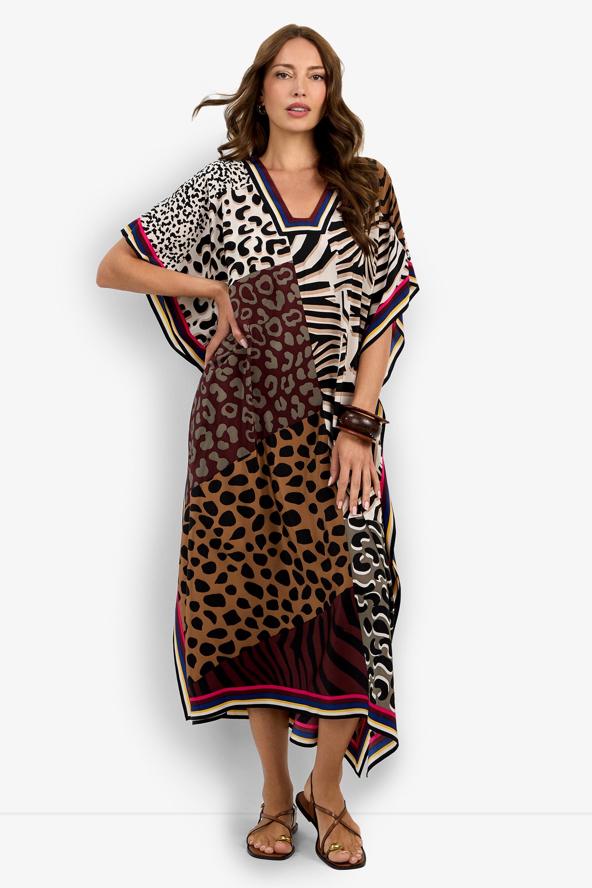 A woman models the Vintage Animal Caftan Dress, a loose, ankle-length piece featuring mixed animal prints and bold hem stripes, styled with brown sandals and chunky bracelets against a white background.