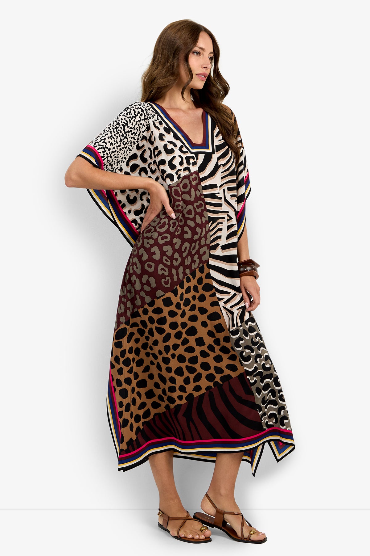 A woman models the Vintage Animal Caftan Dress, a loose, colorful gown with mixed animal prints and pink trim, styled with brown sandals and bracelets.