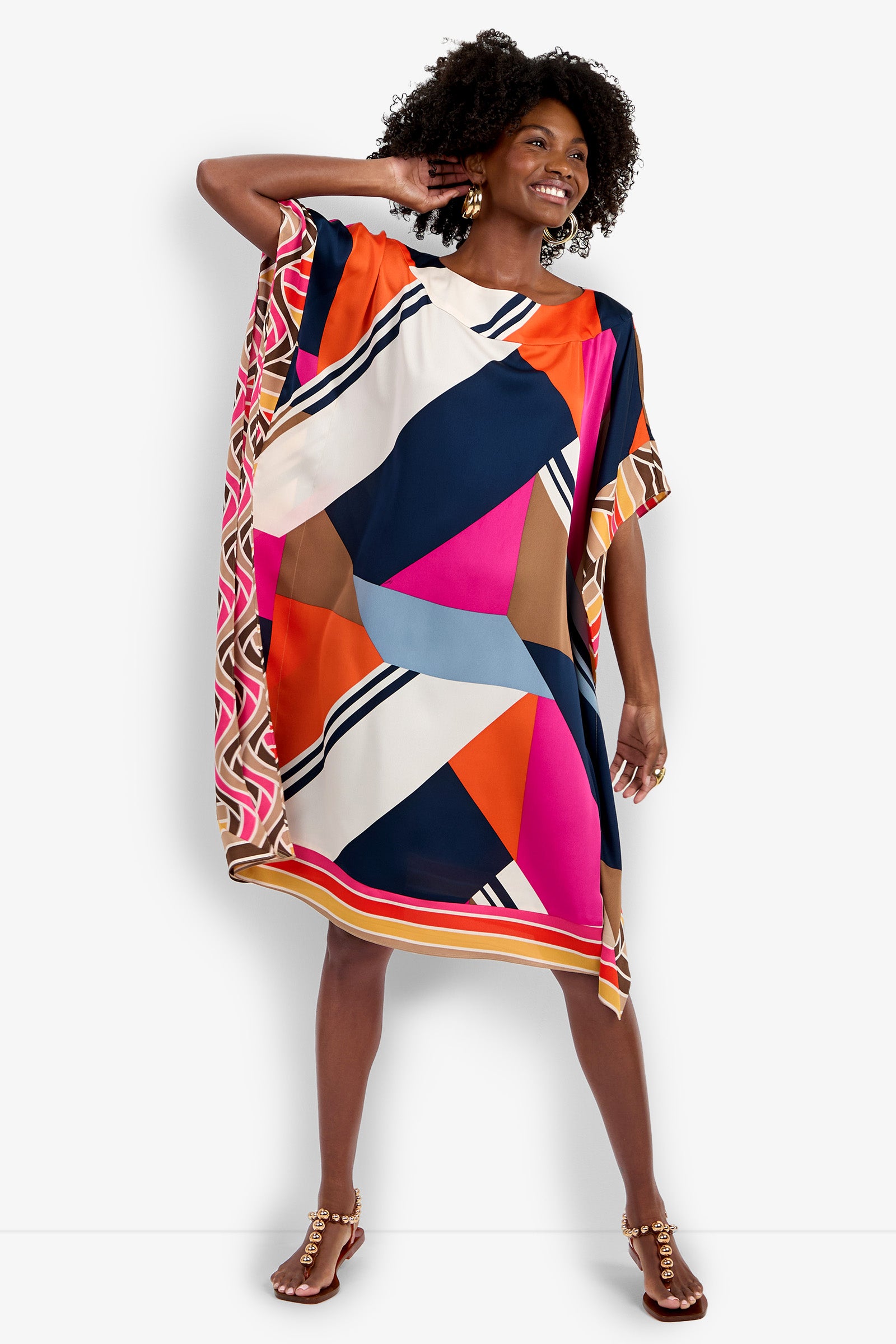 A woman smiles wearing the Vintage Geo Caftan, a loose knee-length dress with bold geometric patterns in navy, pink, orange, brown, and white. The short-sleeve dress features colorful borders and is styled with earrings and sandals.