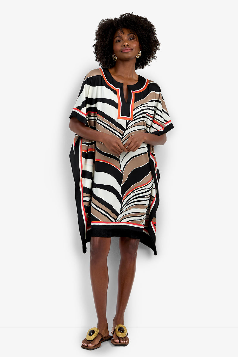 A woman with curly hair and large earrings wears the Vintage Zebra Caftan, a loose, short-sleeved dress featuring an abstract black, white, beige, and red pattern. She pairs it with flat sandals accented in gold.