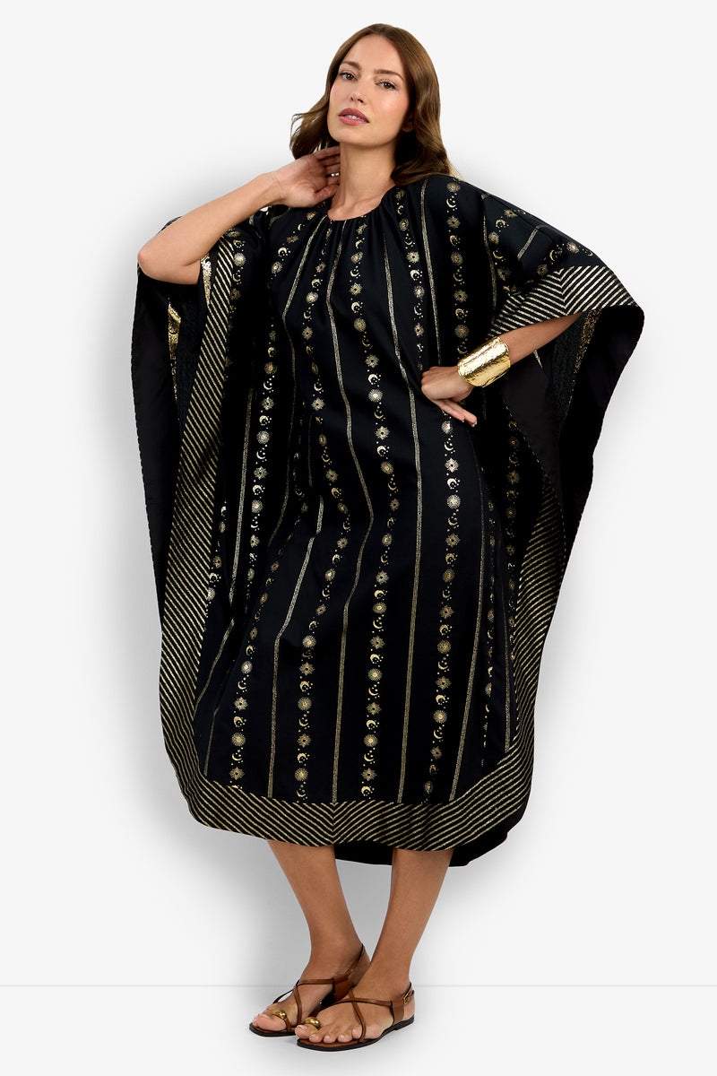 A woman models the Vintage Celestial Caftan, a loose-fitting black dress with gold vertical patterns and a striped gold border. She pairs it with sandals and gold bangles, posing confidently with one hand on her hip.
