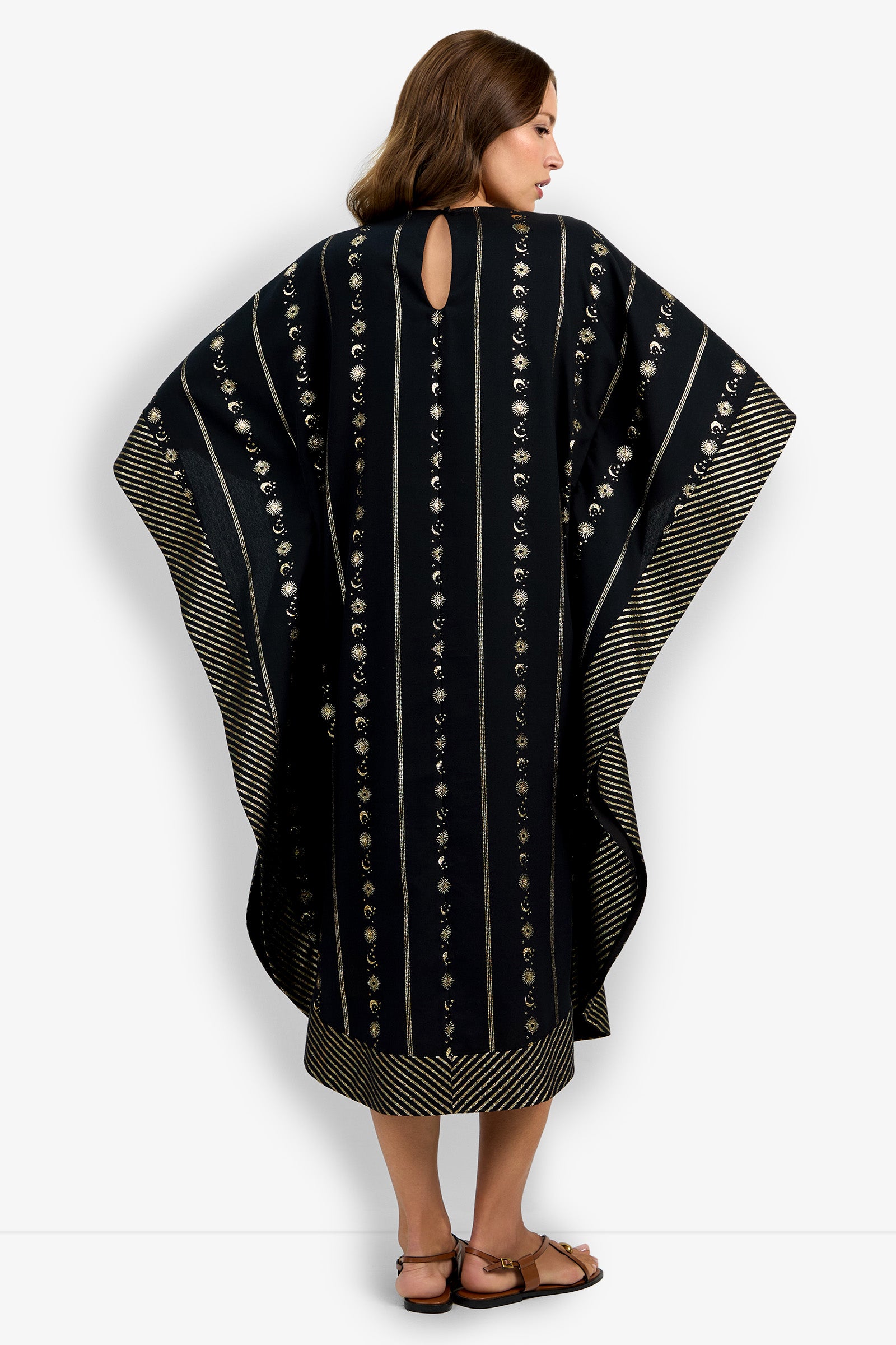 A woman with brown hair and sandals stands facing away, wearing the Vintage Celestial Caftan—a loose black garment featuring vertical gold and silver embroidery and a striped border.