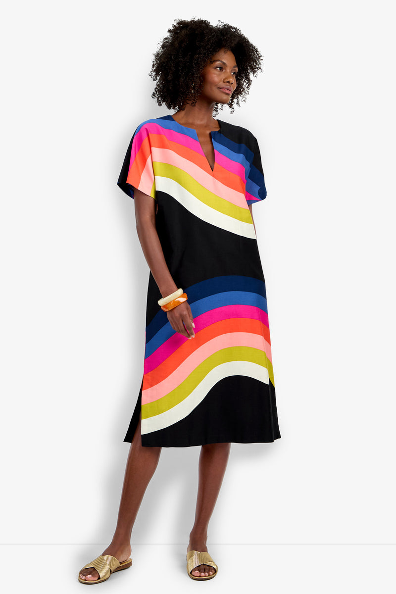 A woman with curly hair and gold bracelets wears the Vintage Rainbow Caftan, a knee-length black dress featuring colorful wavy stripes in pink, orange, yellow, white, and blue. She completes the look with beige slide sandals.