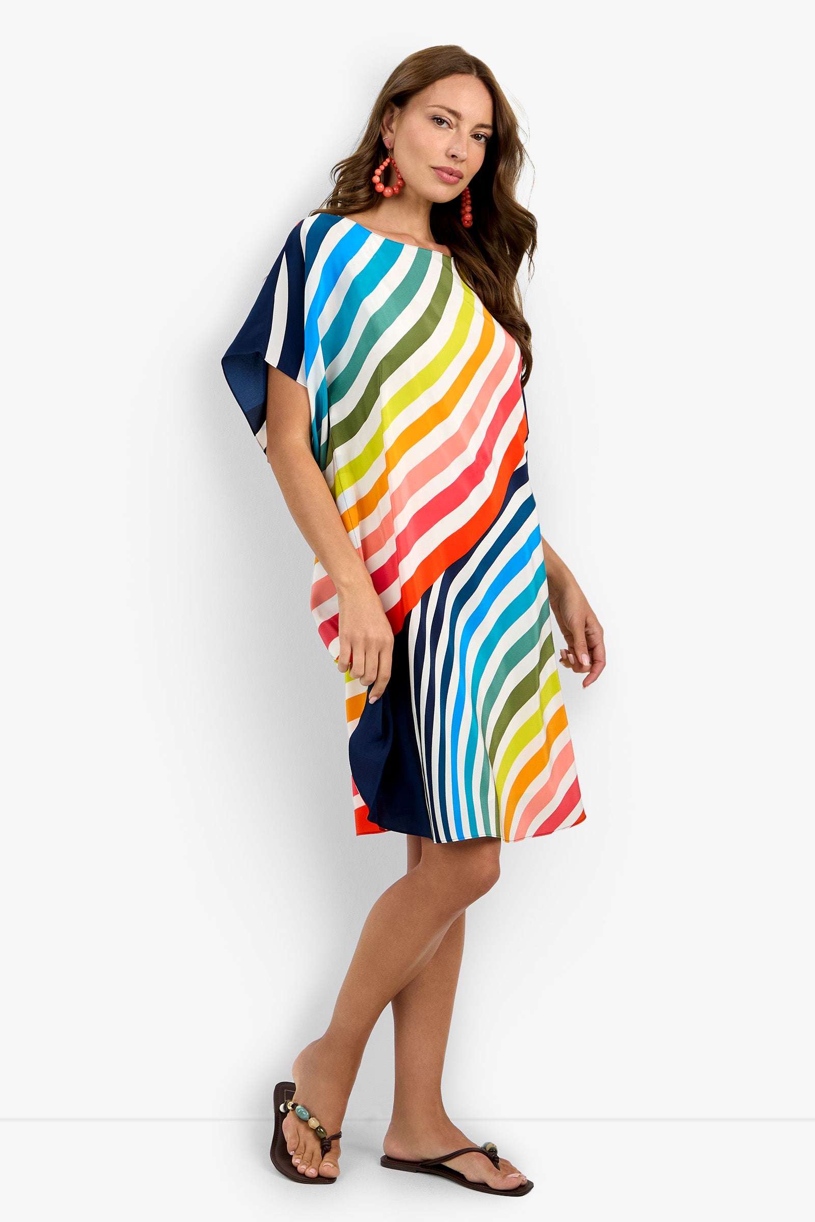 A woman in the Vintage Ombre Rainbow Caftan—a loose, knee-length dress with colorful diagonal stripes and short sleeves—pairs it with black sandals and red earrings, standing against a plain white background.