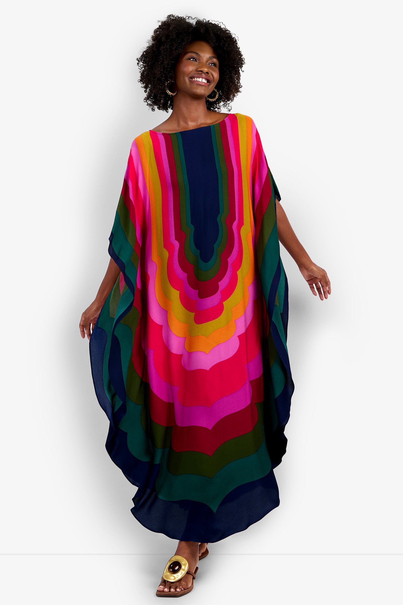 A smiling woman with curly hair wears the Vintage Moroccan Geo Caftan, featuring bold, wavy stripes in pink, red, orange, yellow, green, and blue. Barefoot, she stands against a plain white background.