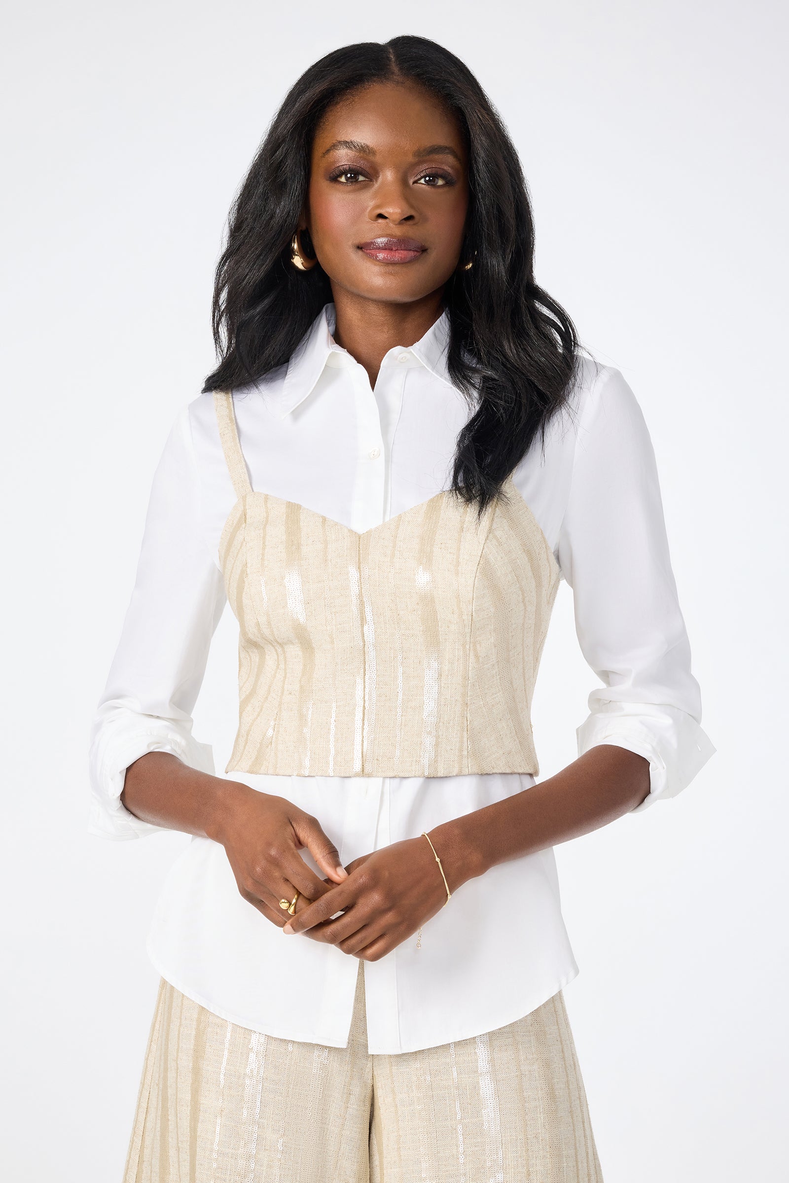 A woman with long dark hair smiles gently in the Millau Sleeveless Top in Biarritz Beige, layered over a white button-up shirt with matching beige pants, standing against a plain white background.